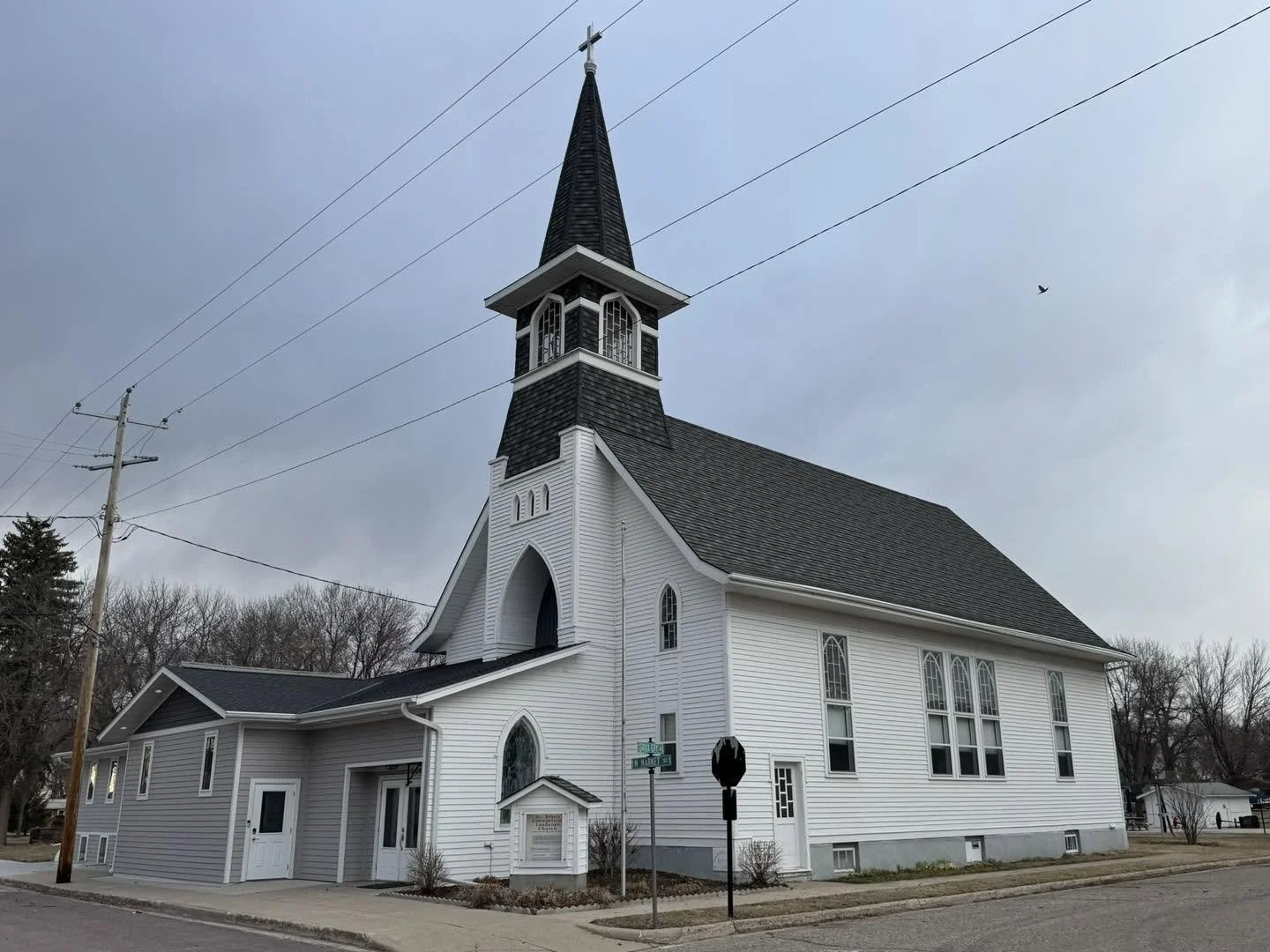 A white church with a tall steeple and large arched windows, located on a corner with street signs, under a cloudy sky.