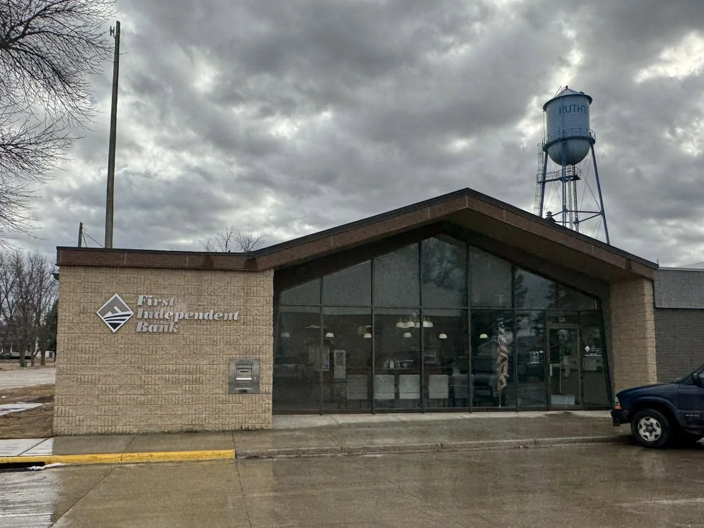 The exterior of a First Independent Bank branch with large glass windows and a brick wall, with a water tower in the background under a cloudy sky