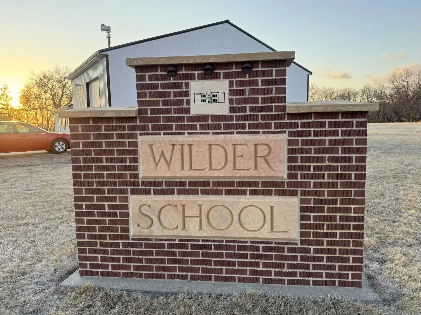 Red brick school sign with the words 'Wilder School' engraved in light-colored stone, in front of a building with a light blue exterior and a sunset in the background.