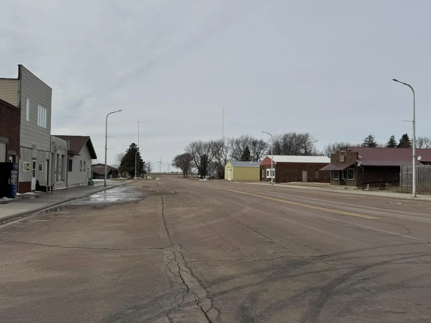 Empty small town street with buildings on the left and right, leafless trees in the background, overcast sky, and wind turbines visible in the distance.