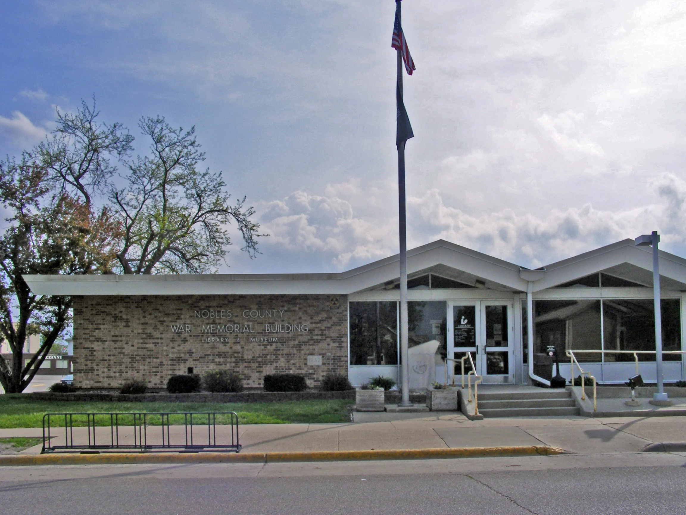 The Nobles County War Memorial Building, which houses a library and museum, with a brick facade, glass entrance, American flag, and flagpole in front. A tree with budding leaves is to the left, and steps lead up to the entrance.