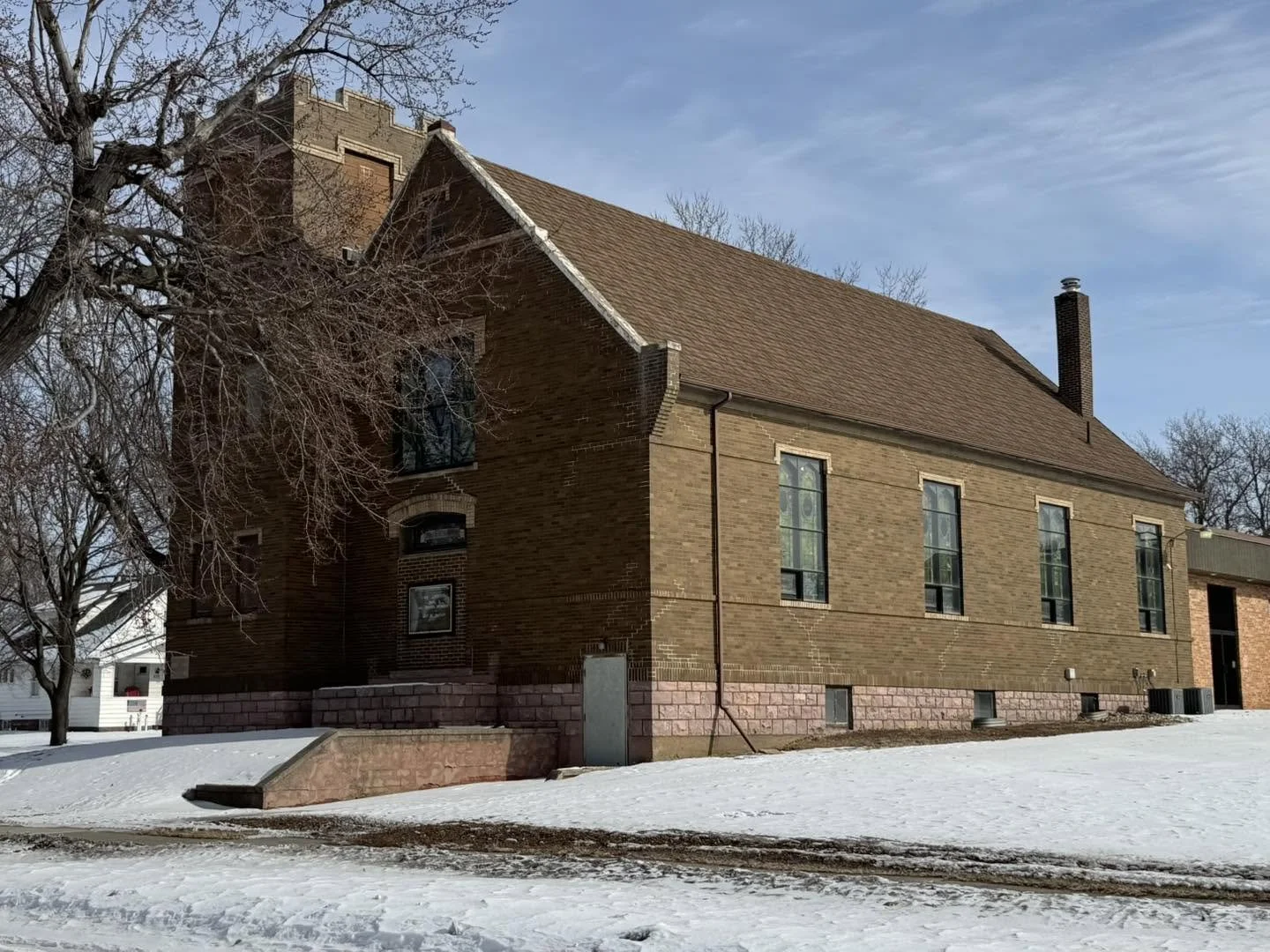 A brick building with tall narrow windows and a high pitched roof, surrounded by snow, with leafless trees and a clear blue sky in the background.