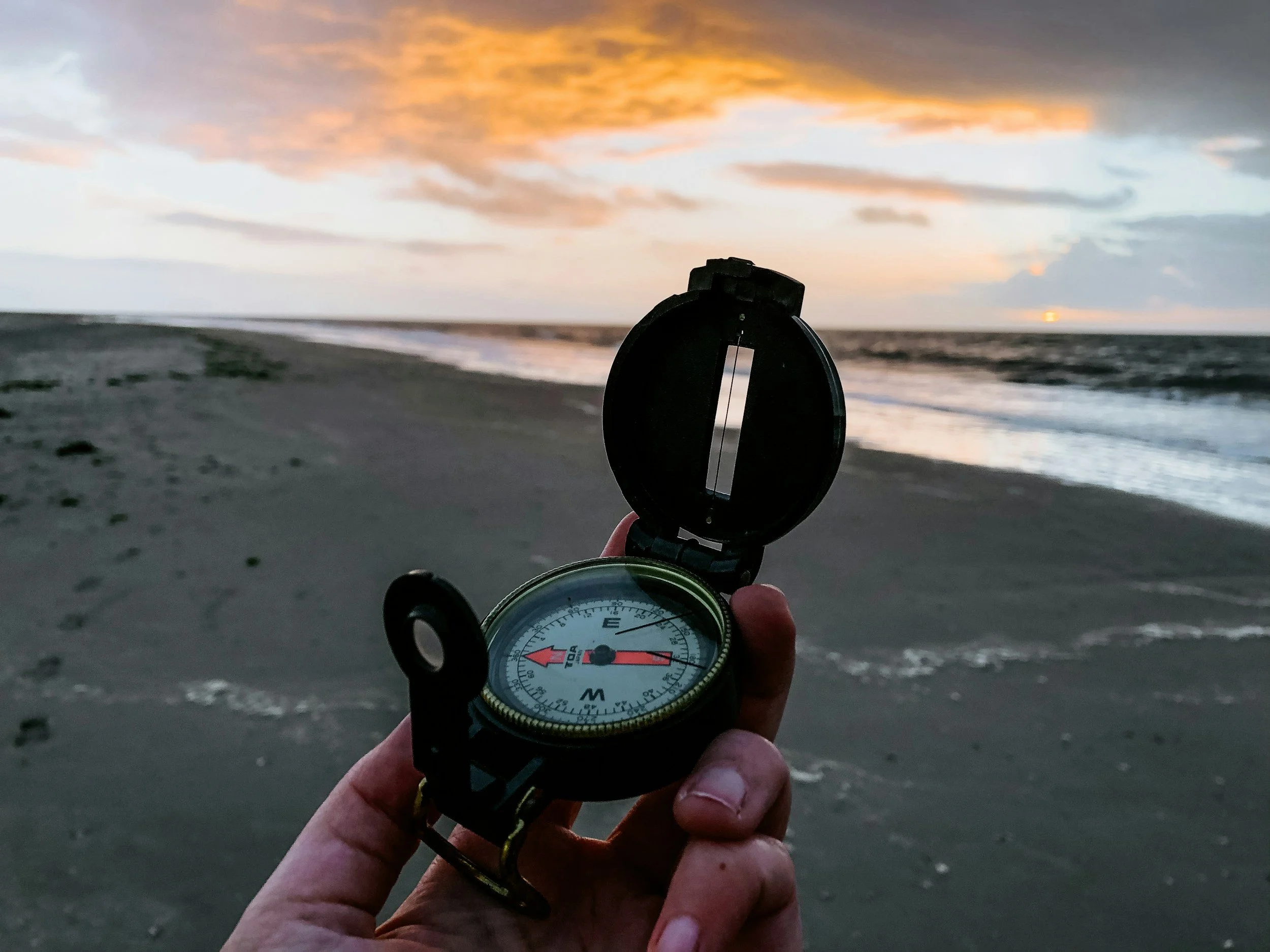 A hand holding a compass on a beach during sunset, with the ocean waves and cloudy sky in the background.