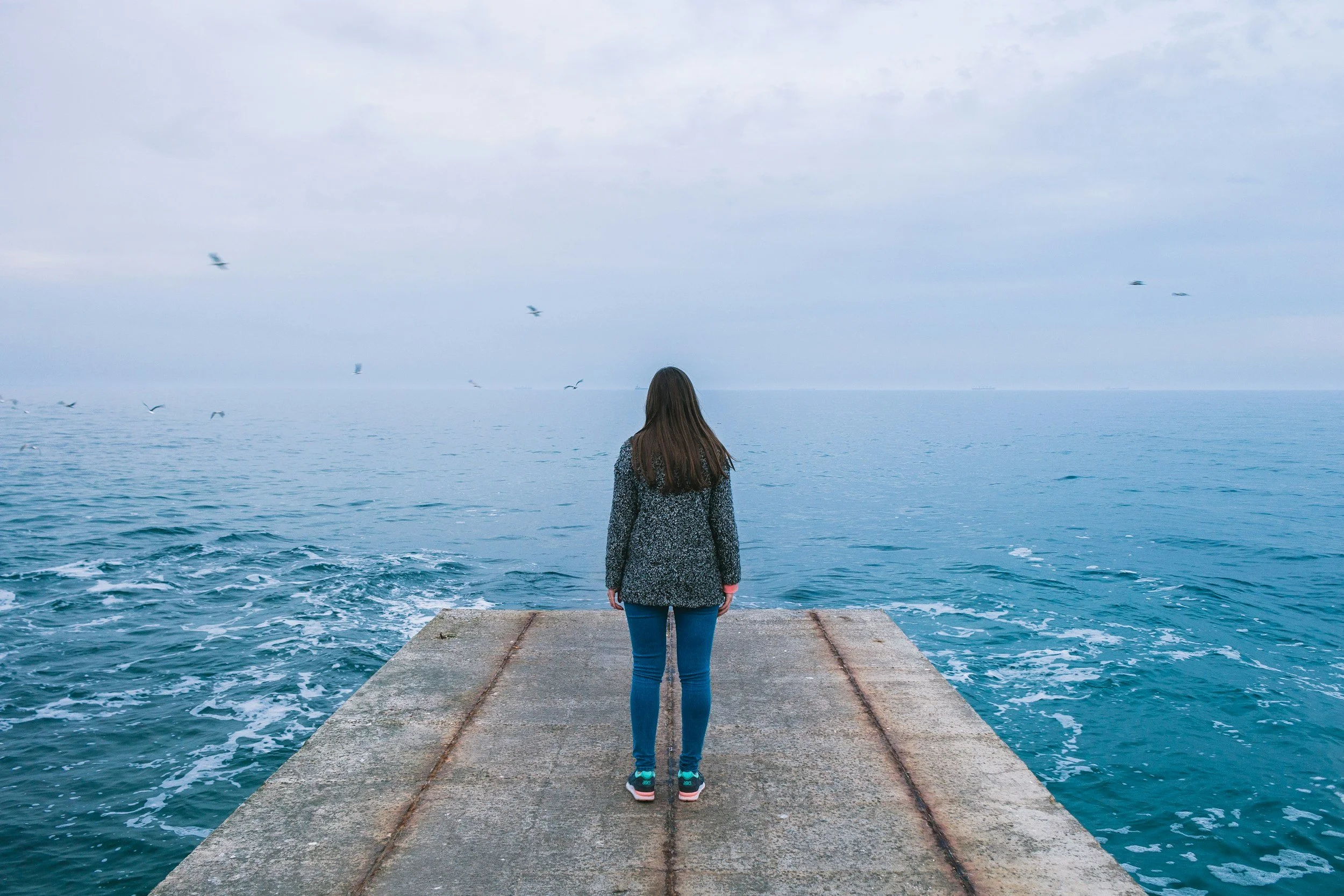 A woman standing on a concrete pier, facing the ocean with her back to the camera. The sky is overcast and seagulls are flying nearby.