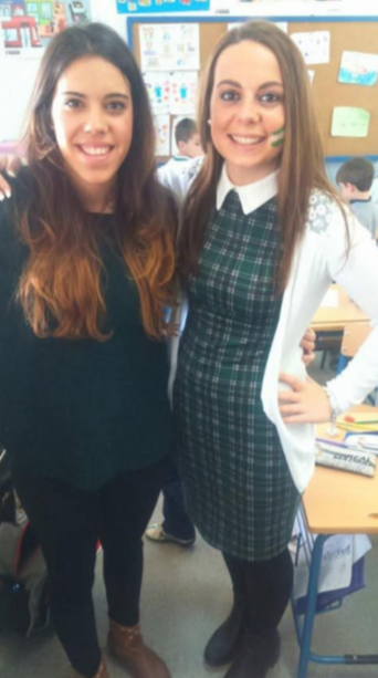 Two young women smiling in a classroom, one wearing a black top and the other in a school uniform with a green plaid dress and white cardigan.