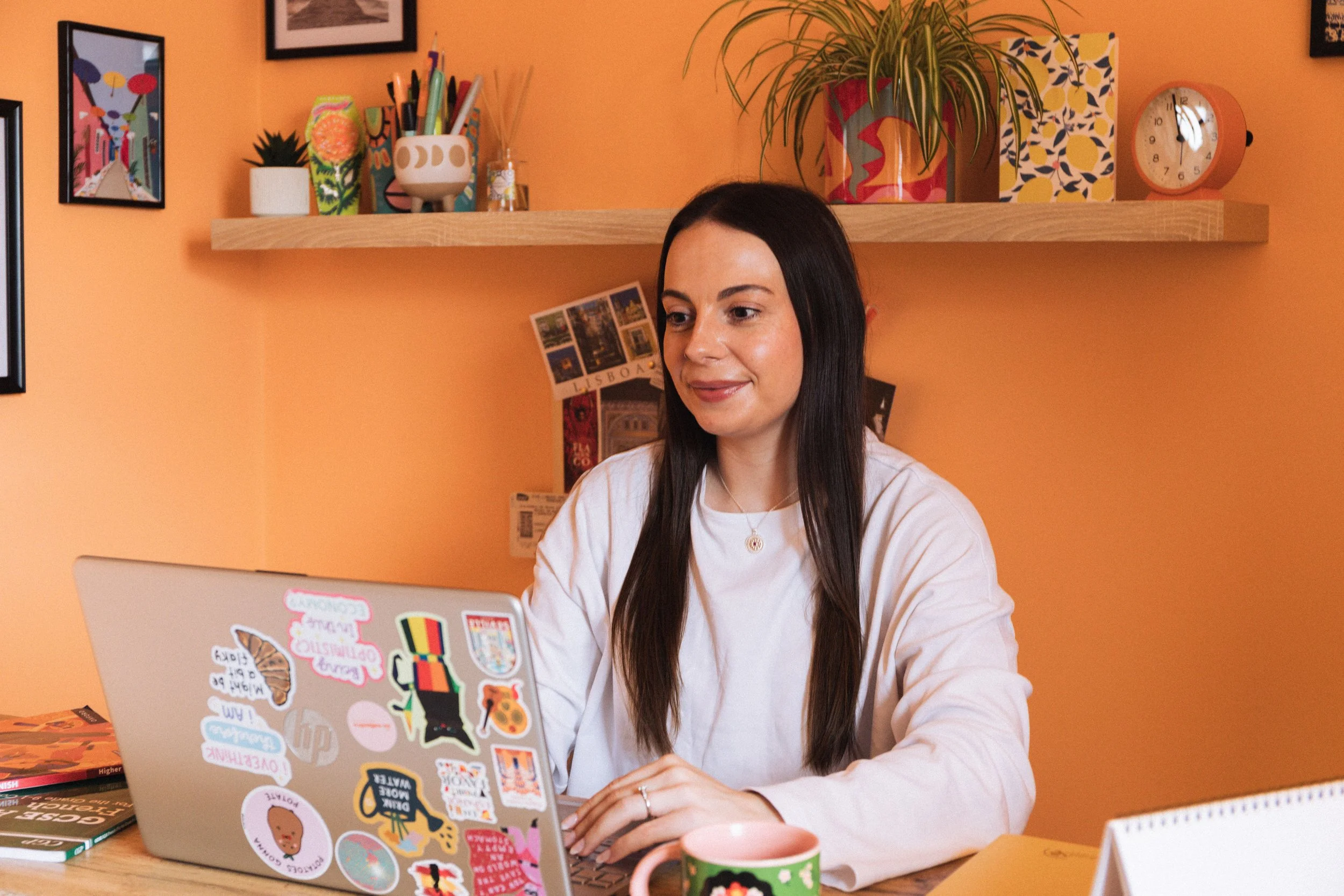 A woman with long dark hair sitting at a desk using a laptop decorated with various colorful stickers, inside a warmly lit room with orange walls and a floating shelf with plants, books, and artwork.