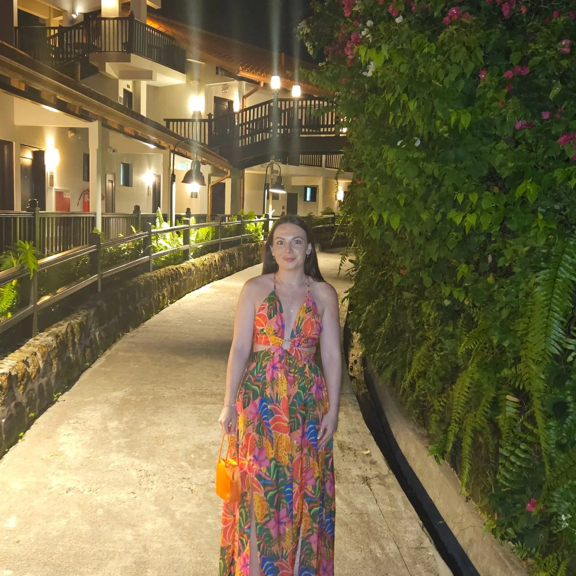 Nighttime scene showing a woman in a colorful, floral-patterned dress standing on a sidewalk next to a green hedge with pink flowers, illuminated by light fixtures against a background of a building with balconies and outdoor lighting.