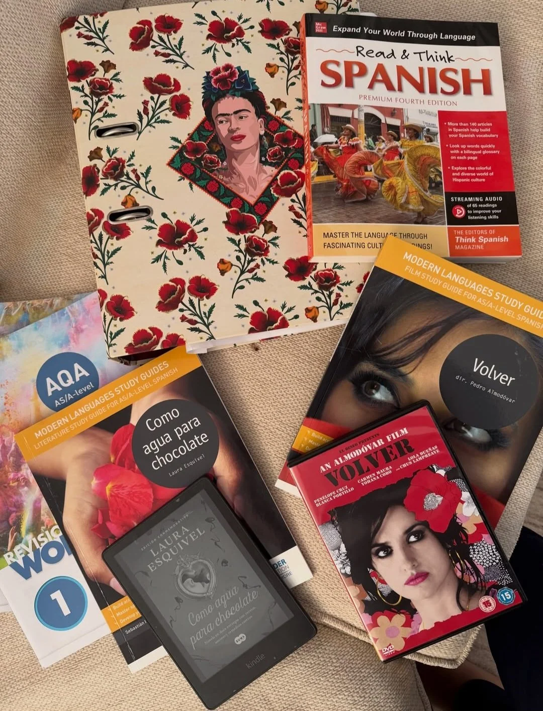 A collection of Spanish language learning materials including books, a DVD, and an e-reader. The books feature titles such as "Read & Think Spanish," "Como agua para chocolate," "Volver," and "Revisión Word 1." The DVD is titled "Wolfver" and shows a woman with dark hair and makeup. The e-reader displays the title "Como agua para chocolate" by Laura Esquivel. There is also a decorative notebook with a floral pattern and an illustration of Frida Kahlo.
