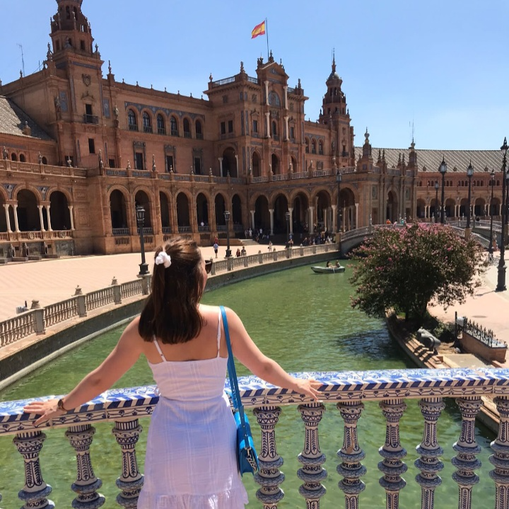 A woman in a white dress and blue purse standing on a decorative bridge overlooking a green pool and the historic Plaza de España in Seville, Spain, with a large building and clear blue sky in the background.