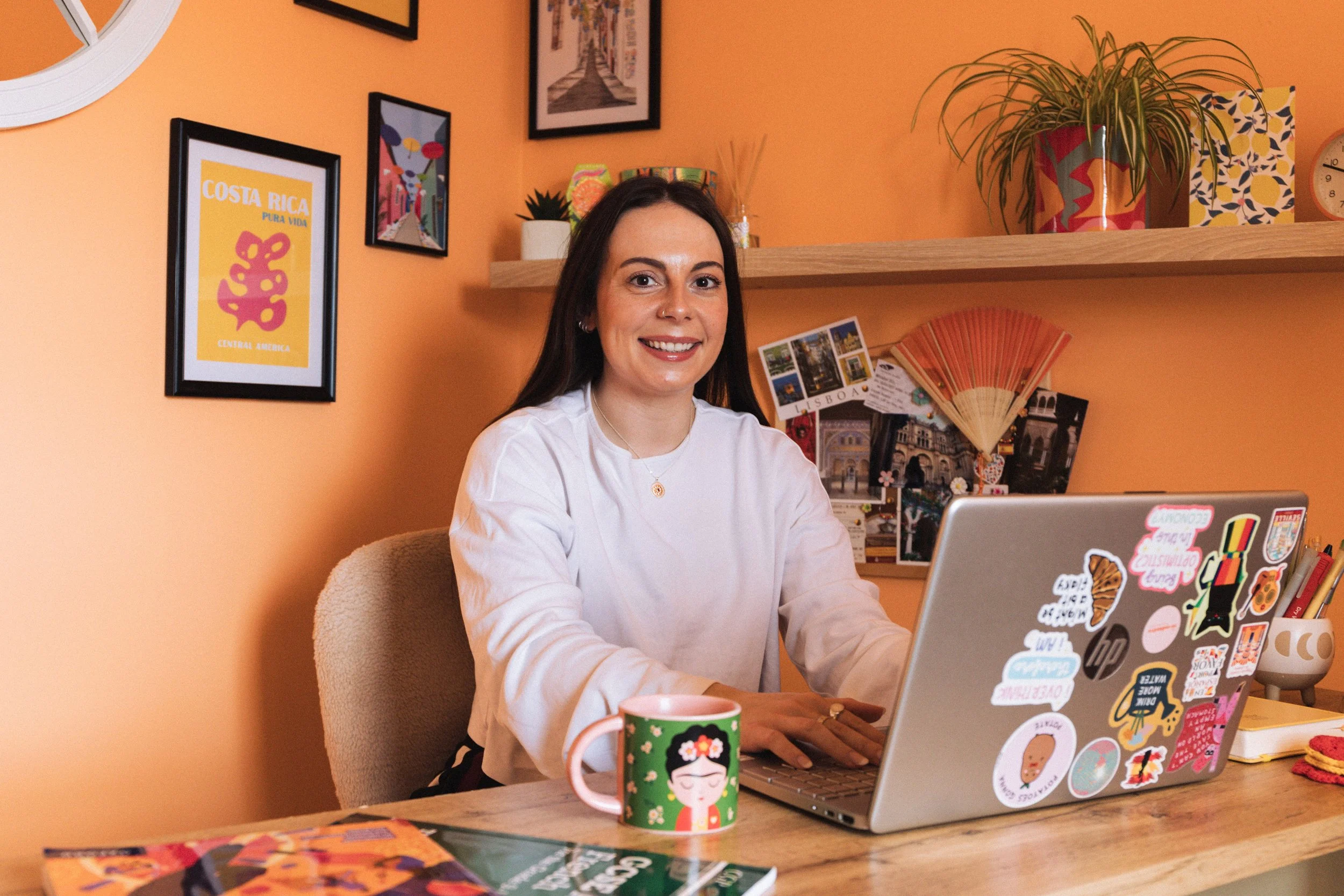 A woman with dark hair and a white shirt sitting at a desk with a laptop covered in colorful stickers, a green mug with a geisha design, and various colorful artworks and posters on the orange wall behind her.