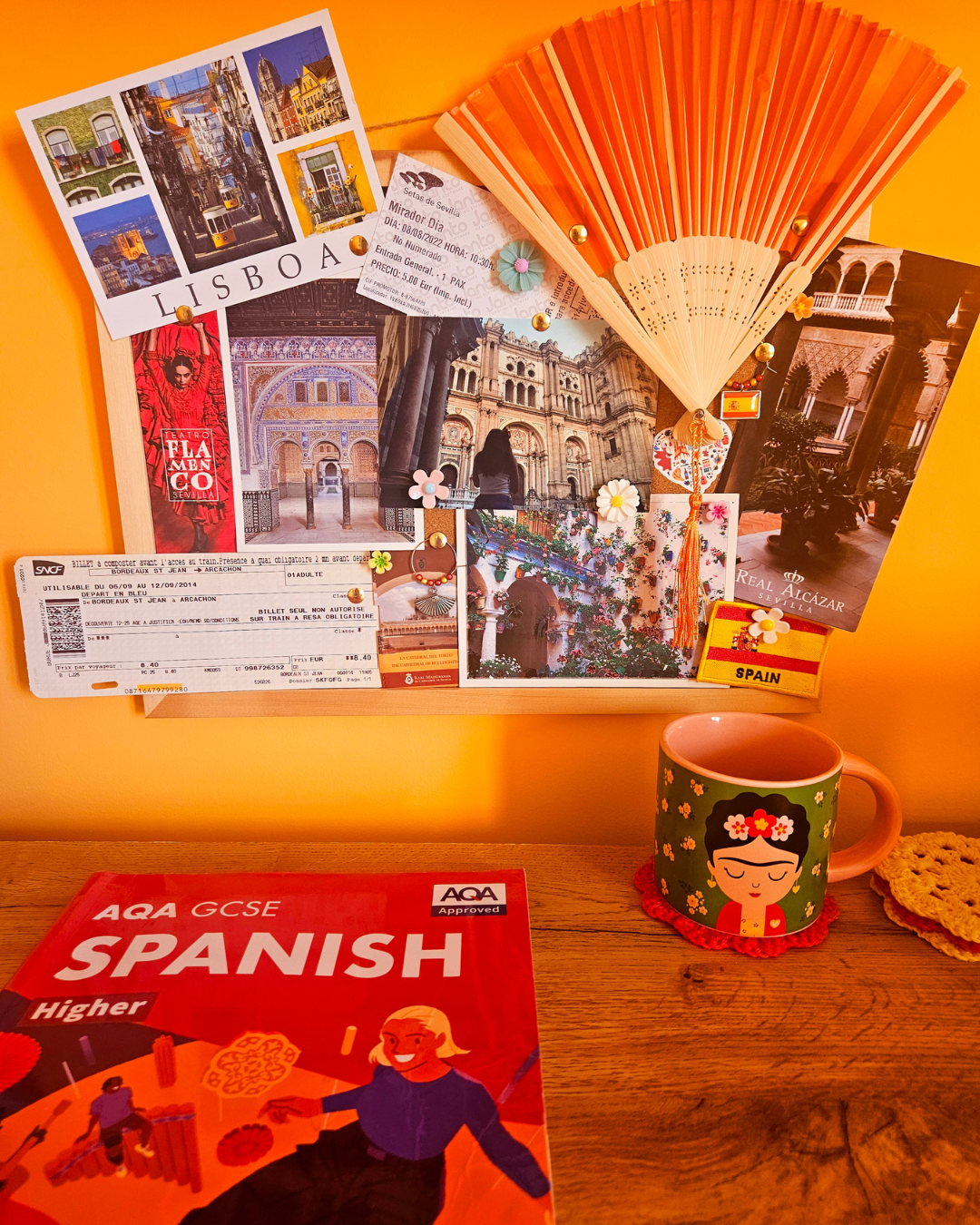 A bulletin board with travel photos and tickets from Lisbon, Portugal and Seville, Spain, decorated with flowers, a fan, and small souvenirs. On the table below, there is a Spanish language book and a mug with a woman and floral design.