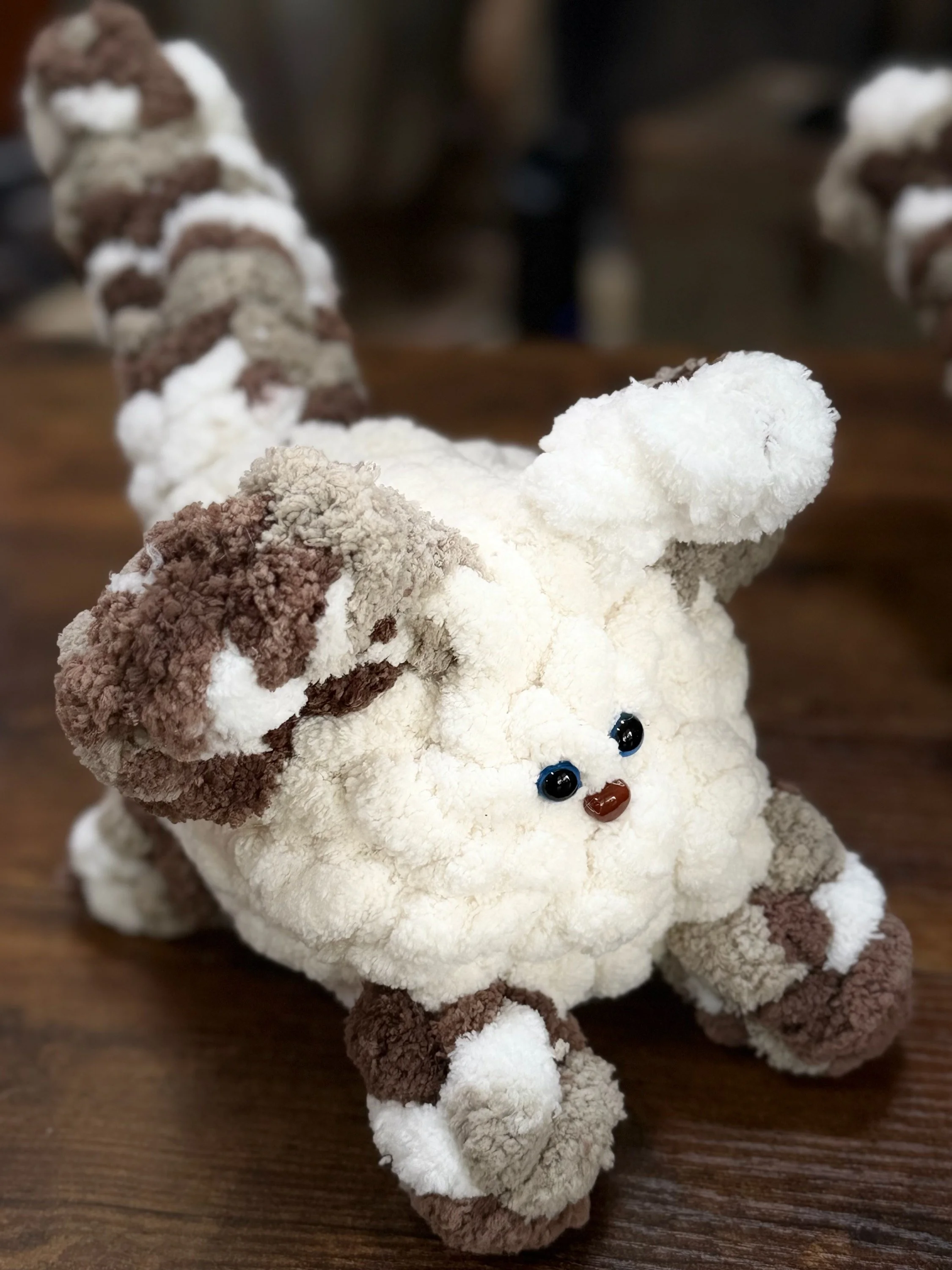 A plush toy resembling a puppy with a multicolored, fluffy, curly fur pattern, blue eyes, and a small brown nose, sitting on a wooden surface.