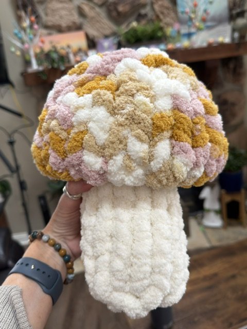 A fuzzy, plush mushroom toy with a white stem and a colorful, fluffy cap in pastel pink, yellow, and white, held by a hand.