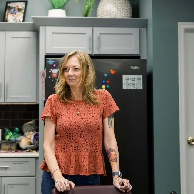 A woman with blonde hair wearing an orange lace top standing in a kitchen with gray cabinets and a black refrigerator.