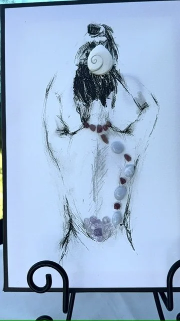 A drawing of a woman with a shell over her face and large shells on her body, with rocks and shells placed over her abdomen, creating an abstract art piece.