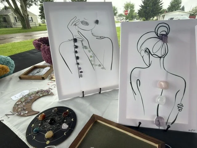 Two framed line art drawings of women with jewelry, displayed on a table outdoors. The left drawing shows a woman with a hand near her face, the right drawing depicts a woman with a bun hairstyle, both accented with semi-precious stones. Various jewelry pieces and stones are arranged on the table.