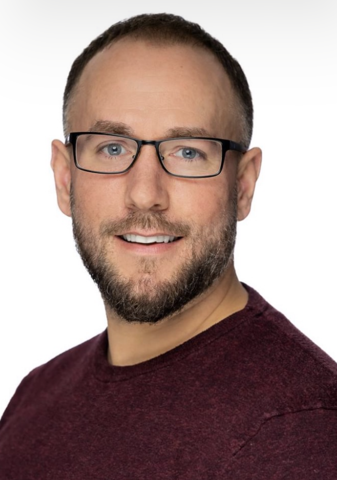 Headshot of a young man with short dark hair, glasses, a beard, wearing a maroon shirt, smiling against a white background.