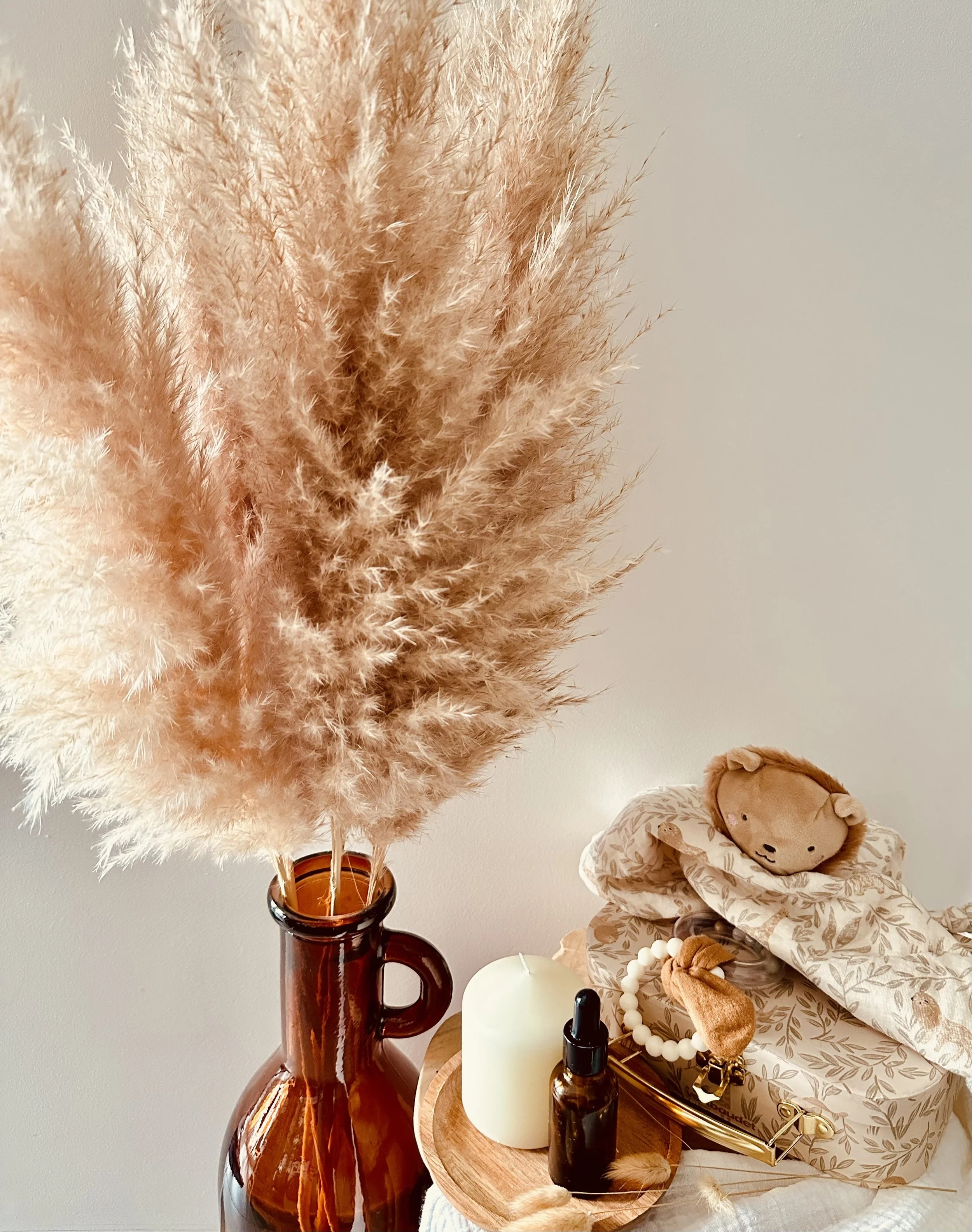 Détails d'une décoration d'intérieur avec un bouquet de fleurs secs en pampa dans un vase en verre brun, une lanterne blanche, des bouteilles en verre, une boîte de bijoux en cuir avec un nounours en peluche et des accessoires de beauté sur une table. Considérant le style, la palette de couleurs est neutre et chaleureuse, avec des éléments en bois et en verre.