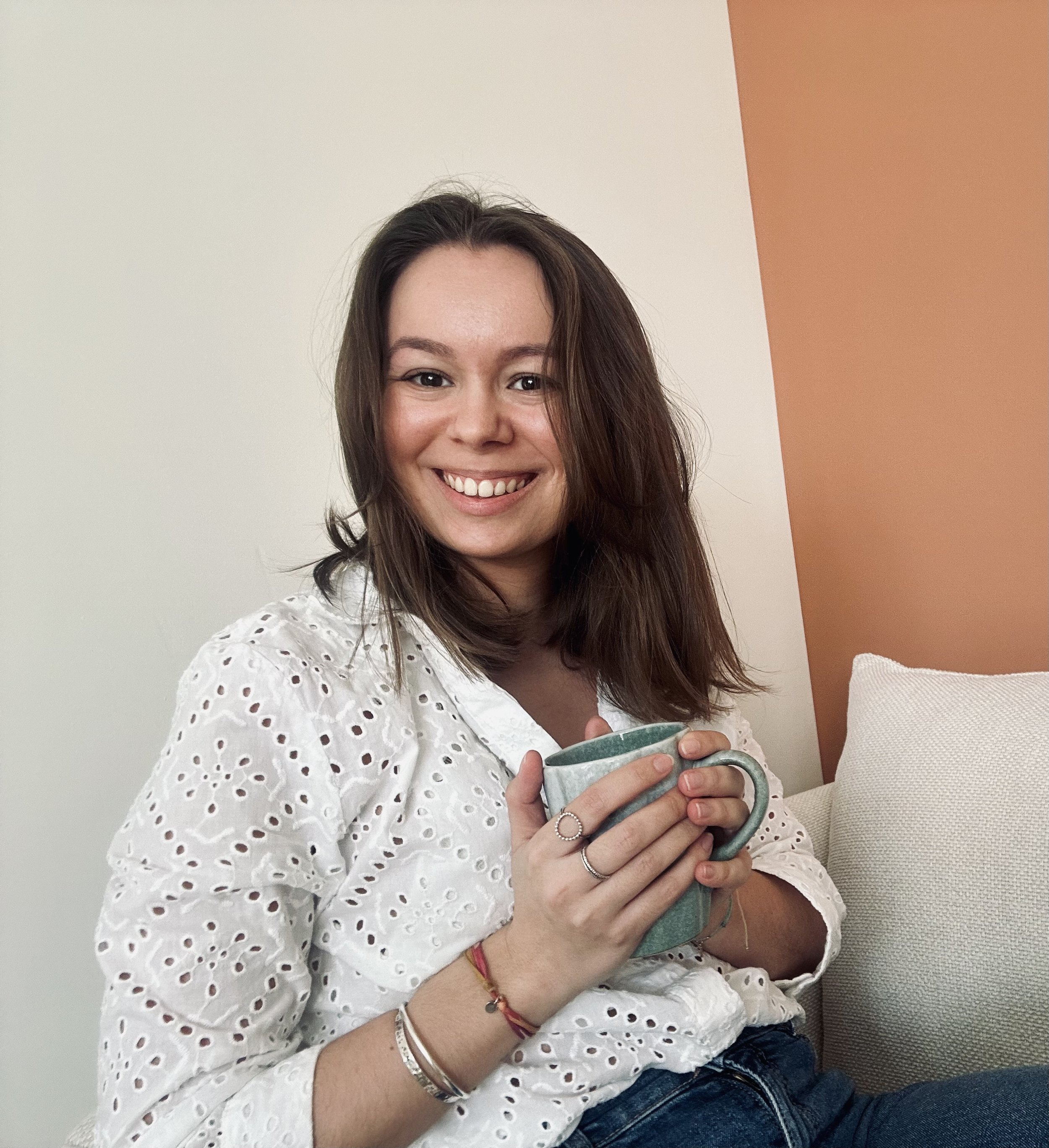 Une femme souriante avec des cheveux bruns, portant une chemise blanche en dentelle, tient une tasse à la main.