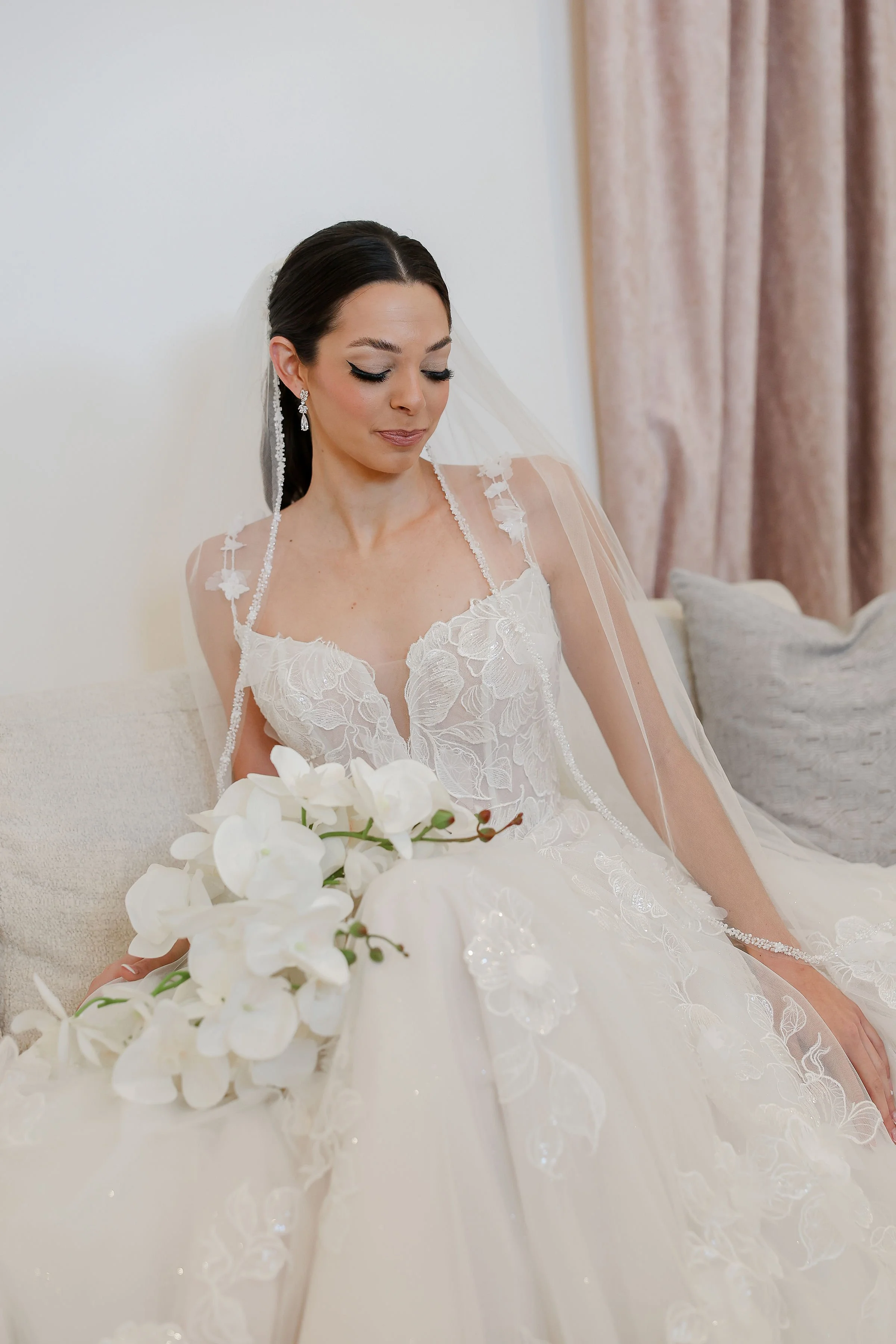 A bride in a white wedding gown with lace details and a veil, sitting on a beige couch, holding a bouquet of white orchids, with beige curtains in the background.