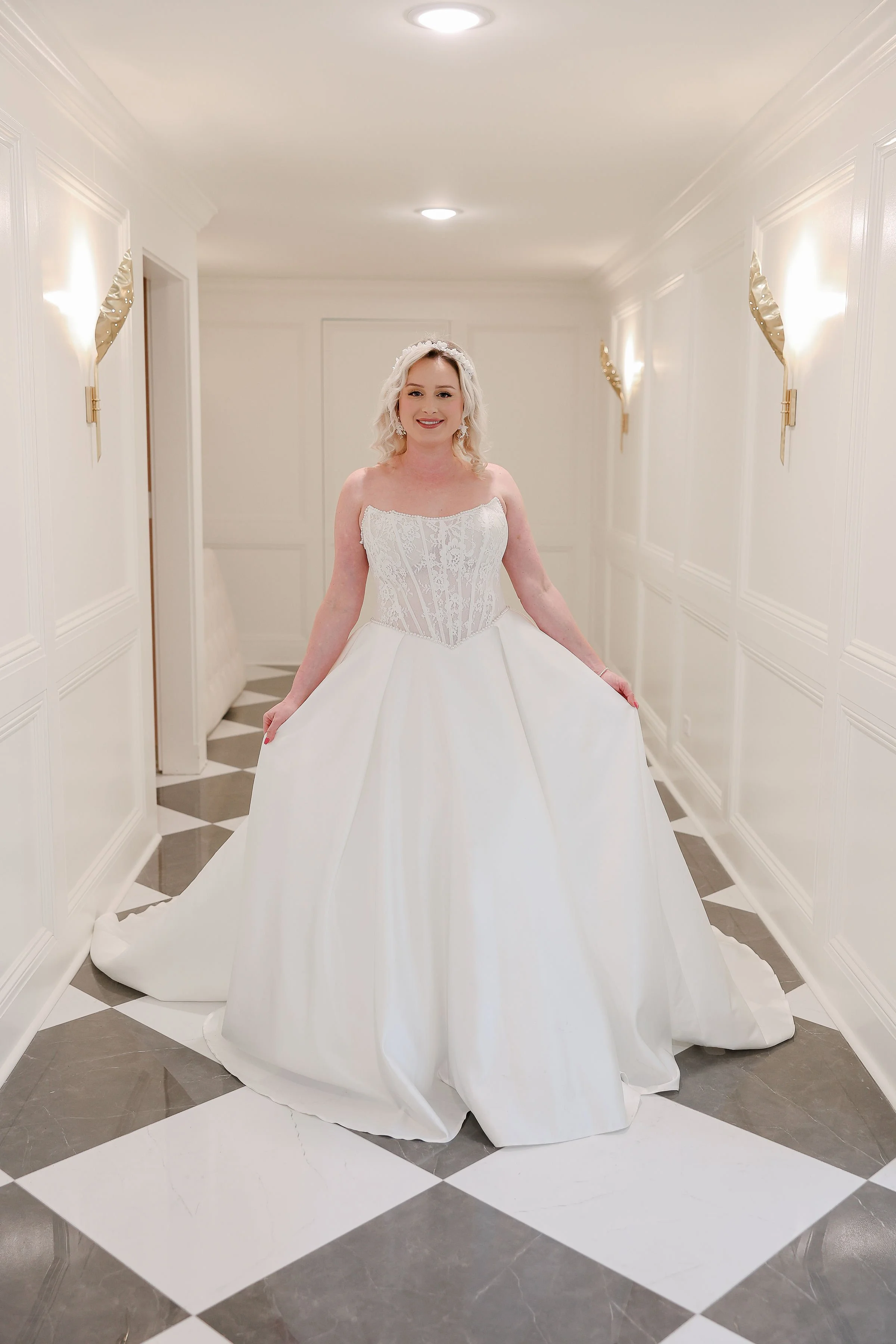 A woman in a white wedding gown standing in a brightly lit hallway with white walls and patterned gray and white floor tiles.