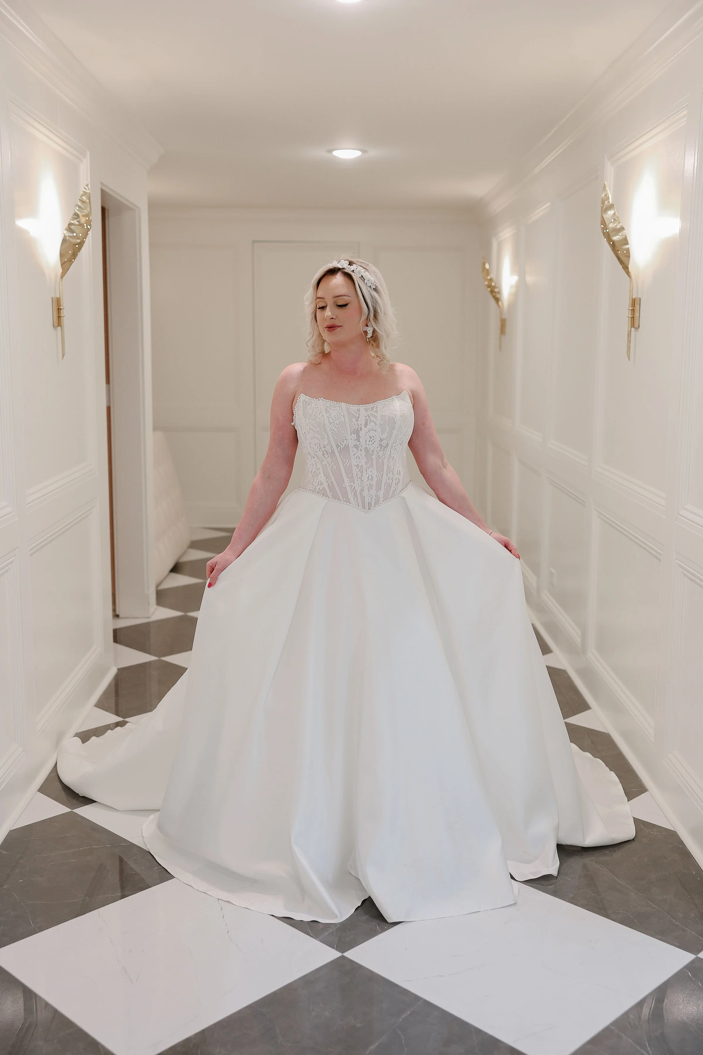 A woman in a white wedding dress standing in a brightly lit hallway with white paneled walls and black and white checkered floor tiles.