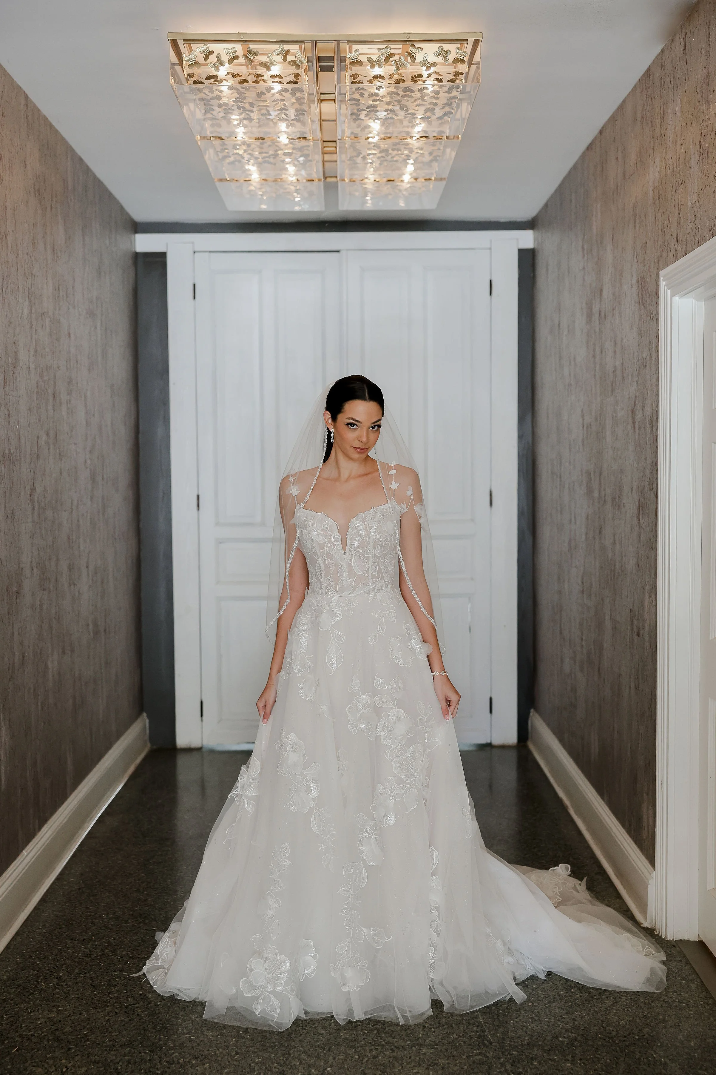 Bride in a white wedding gown with floral embroidery, standing in a hallway with beige walls and white double doors behind her.