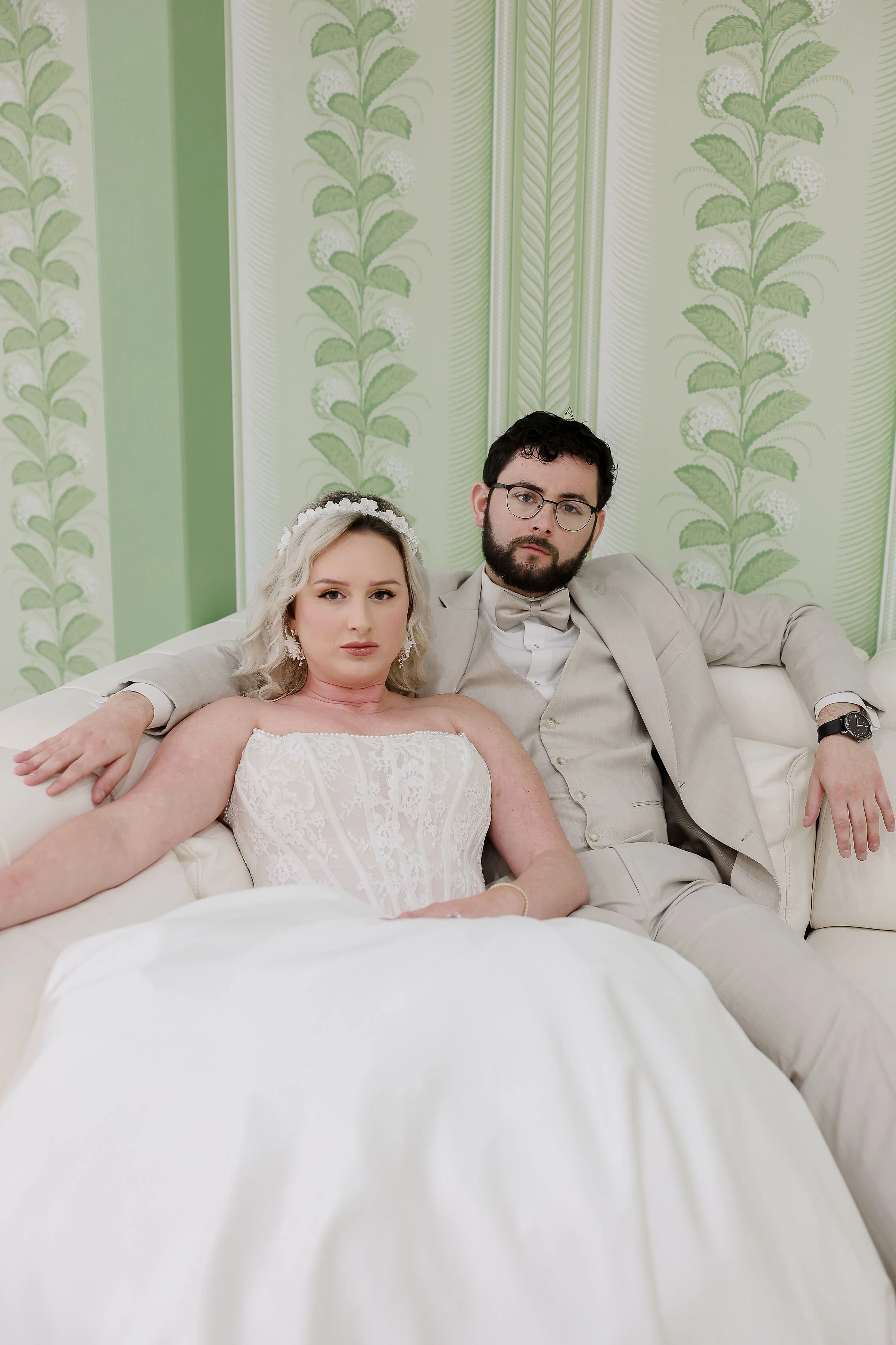 A bride and groom sitting closely on a white couch in a room with green and white floral wallpaper. The bride is wearing a white lace wedding dress and a flower headband, and the groom is wearing a cream suit with a bow tie and glasses.