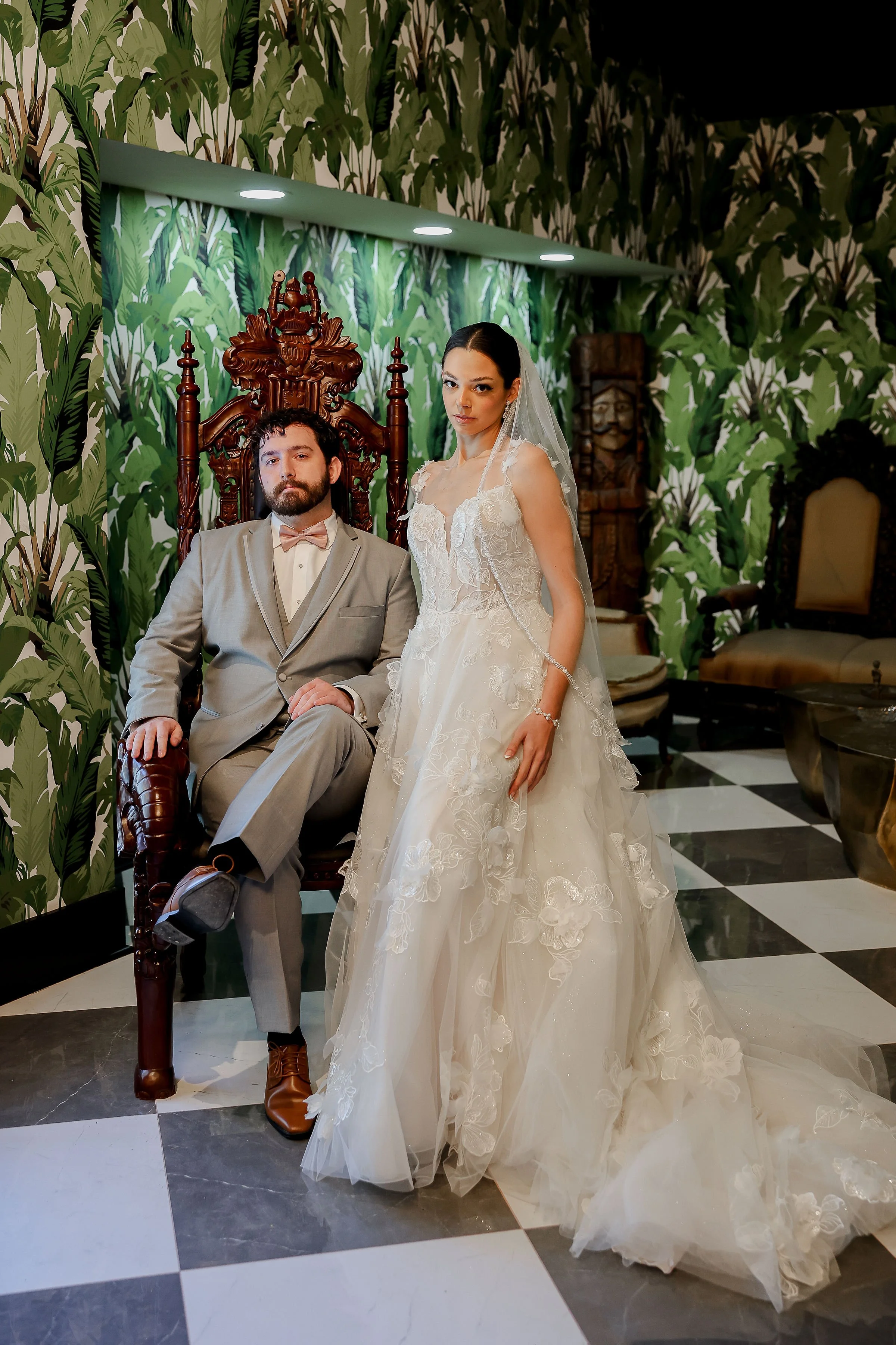 A bride and groom posing indoors; the groom sits on a carved wooden throne-like chair, wearing a gray suit, and the bride stands beside him, wearing a white wedding gown with lace details and a veil. The background has wallpaper with large green leav