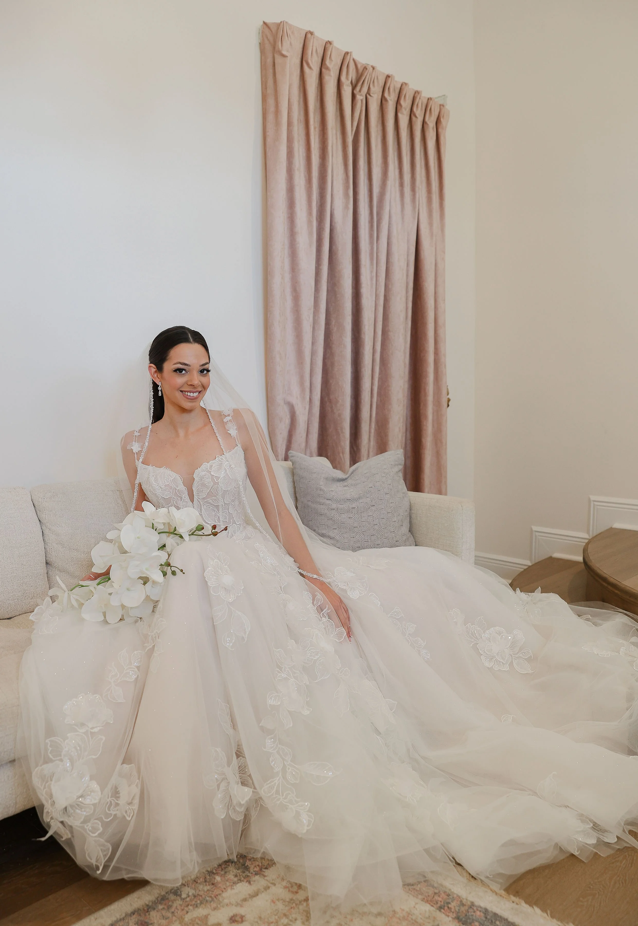 A bride in a white wedding gown sitting on a beige sofa with light gray pillows, holding a bouquet of white orchids, indoors with cream-colored walls and a pink drape background.