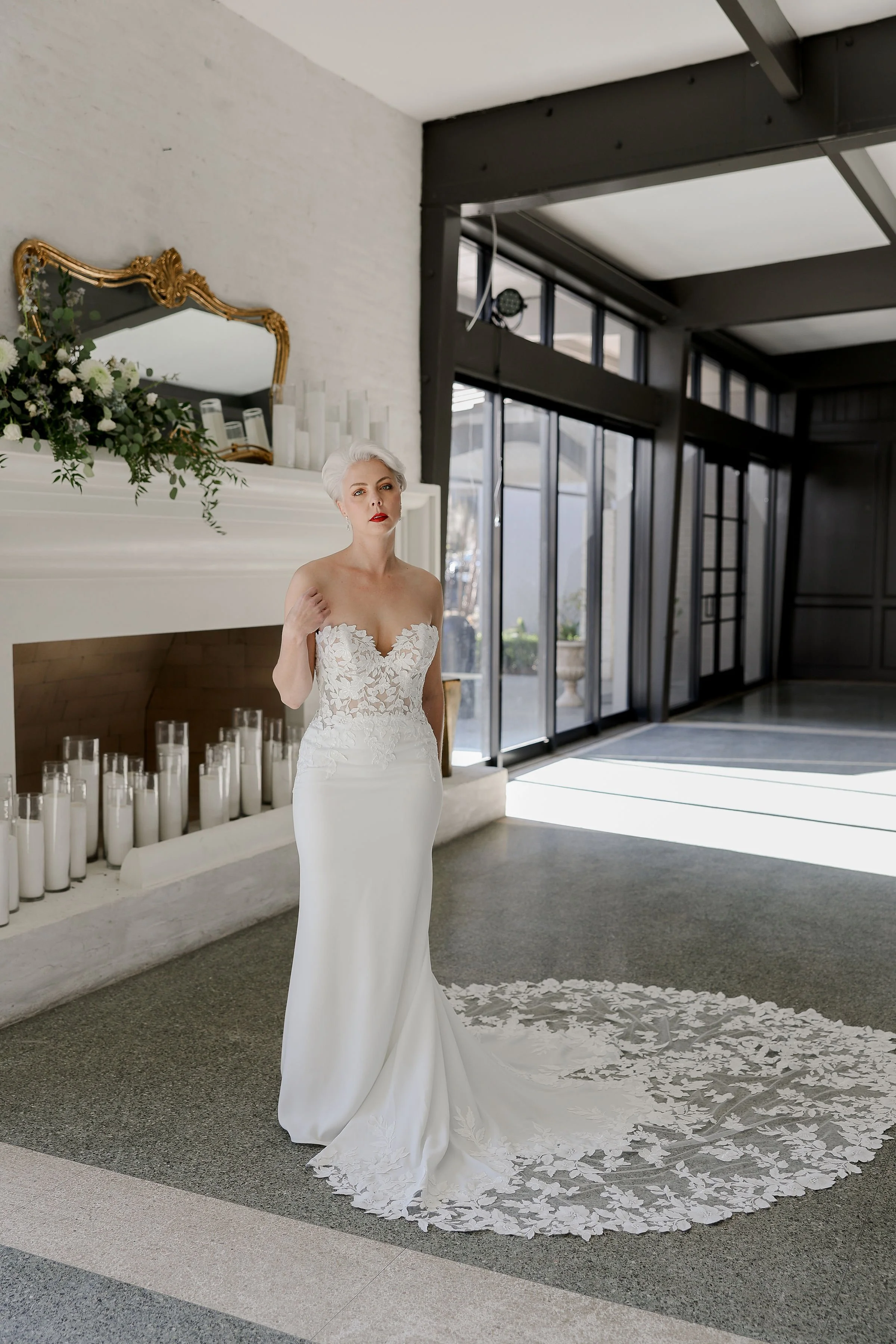 A bride in a white lace wedding gown standing in a modern indoor venue with large windows, candles, and a fireplace decorated with flowers and greenery.