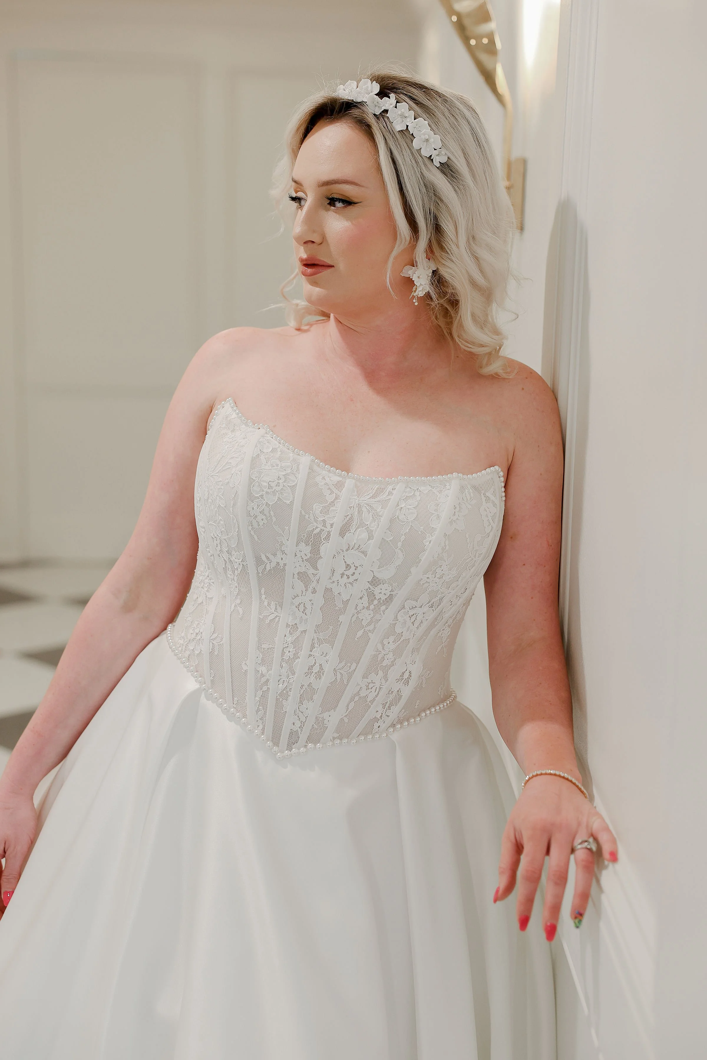 Close-up of a woman in a wedding dress with floral accessories in her blonde hair, standing against a white wall, showing her face, shoulder, hand with rings, and the upper part of her dress.
