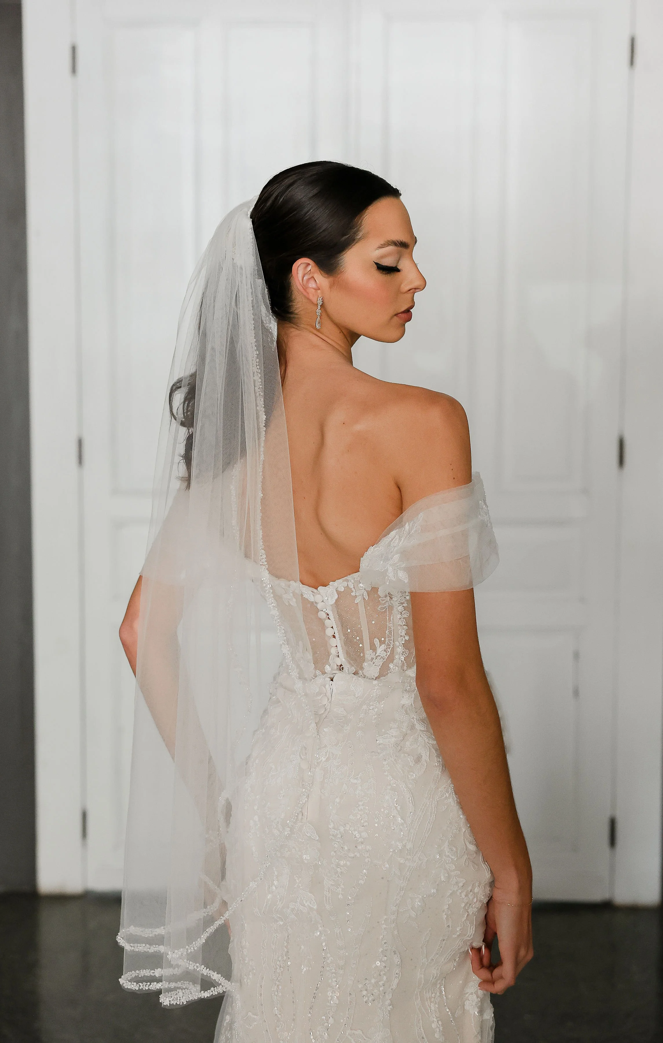 A bride with dark hair in a wedding dress with off-the-shoulder sleeves, wearing earrings and a veil, standing with her eyes closed in front of white paneled doors.