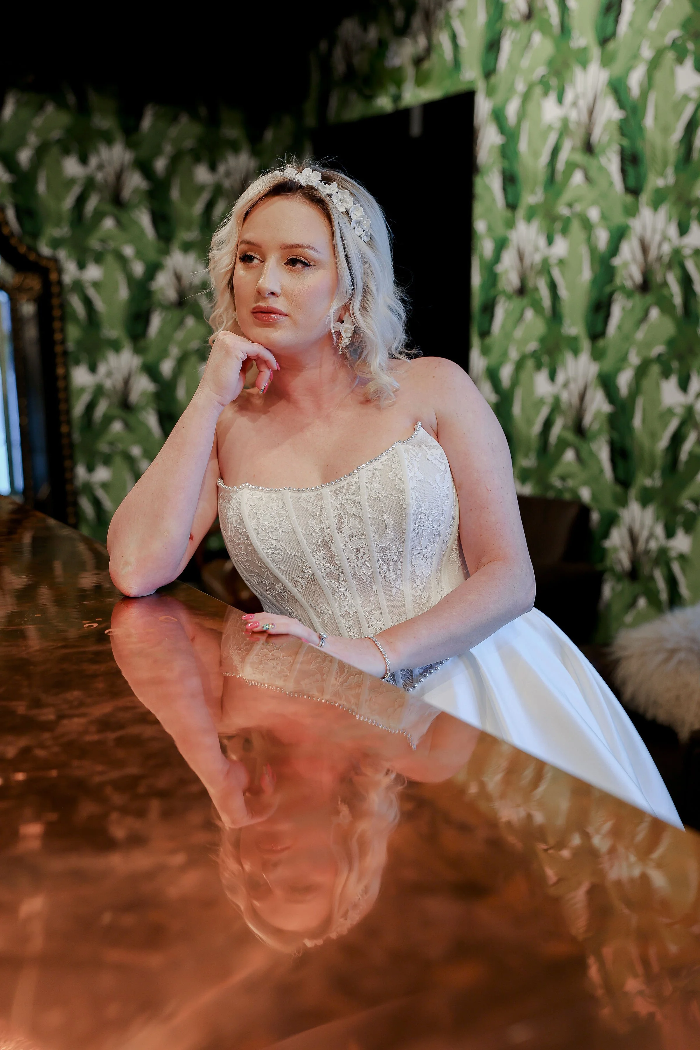 A woman in a wedding dress with a floral headband sitting at a polished wooden table, with her hand resting on her chin, in front of a leafy green wallpaper background.