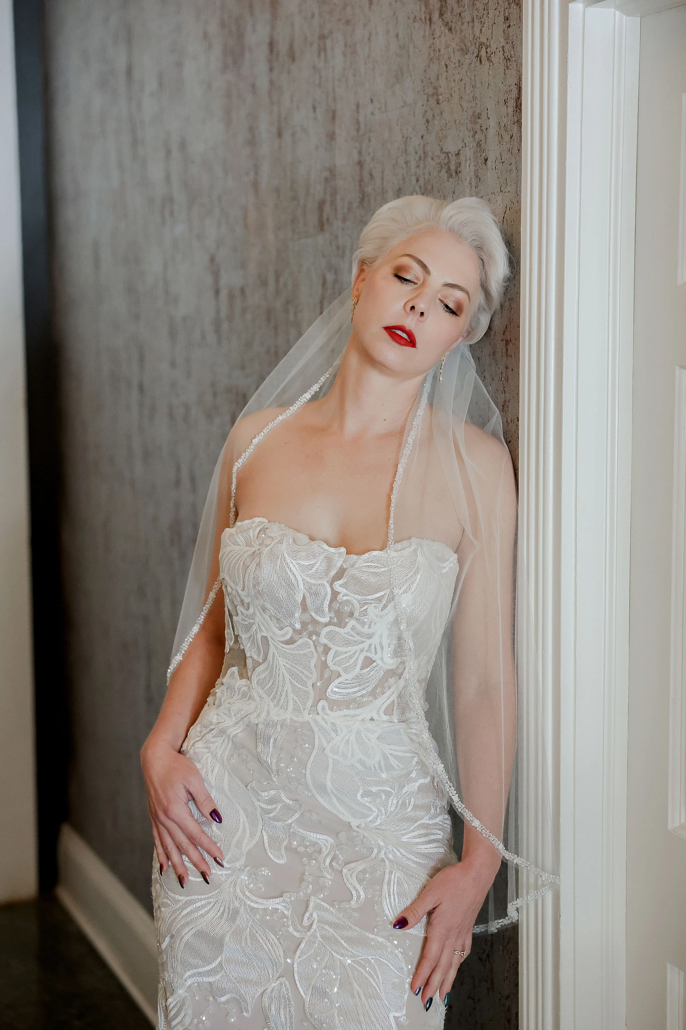 An elderly woman with platinum blonde hair in a wedding dress, leaning her head against a wall with her eyes closed.