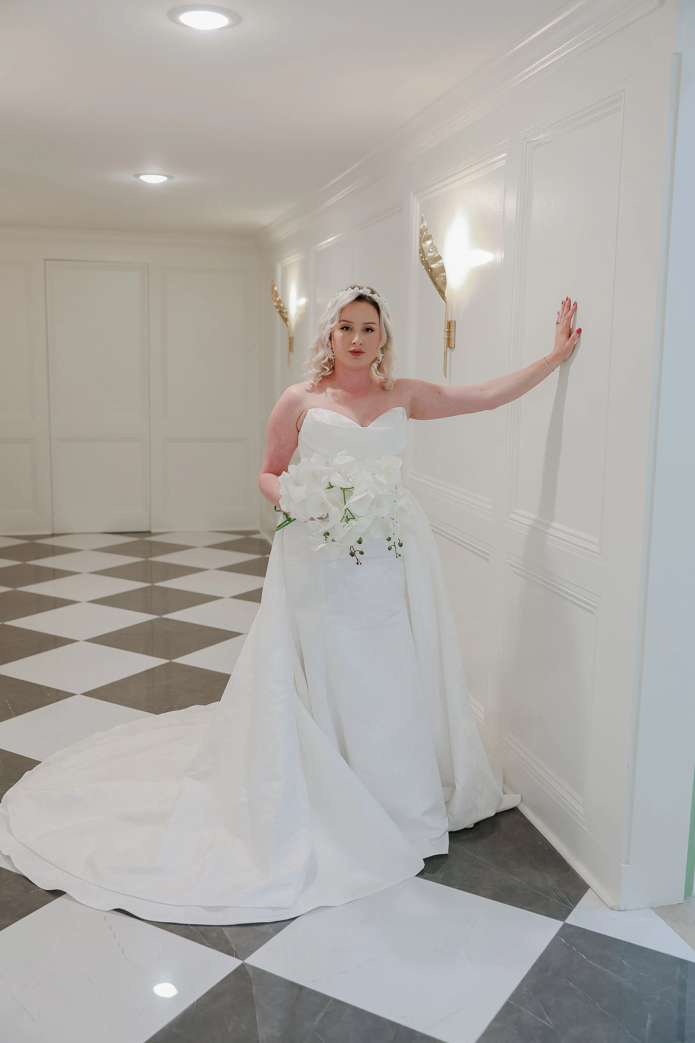 A woman in a white strapless wedding dress holding a bouquet of white flowers, standing in a hallway with white walls and black and white checkered tile floor.