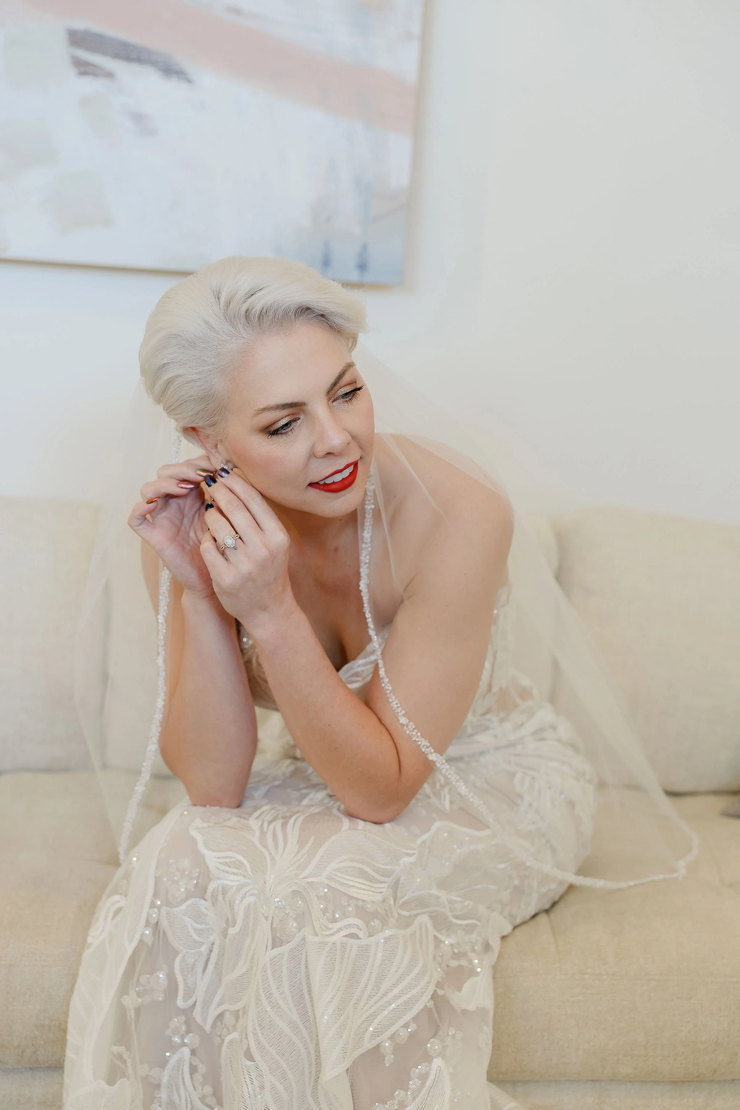 A woman with short platinum blonde hair and red lipstick, wearing a white lace dress with floral embroidery, is sitting on a beige couch, putting on earrings.