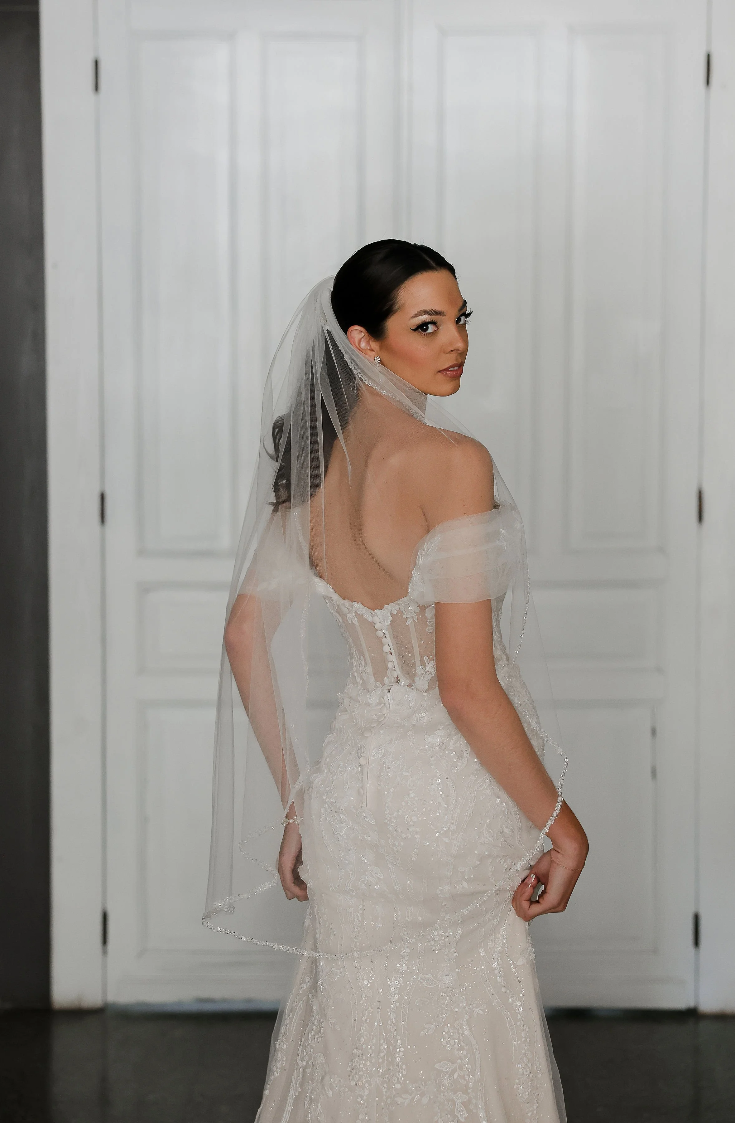 A woman with dark hair, wearing a white wedding dress and veil, turns slightly to look over her shoulder in front of a white paneled door.