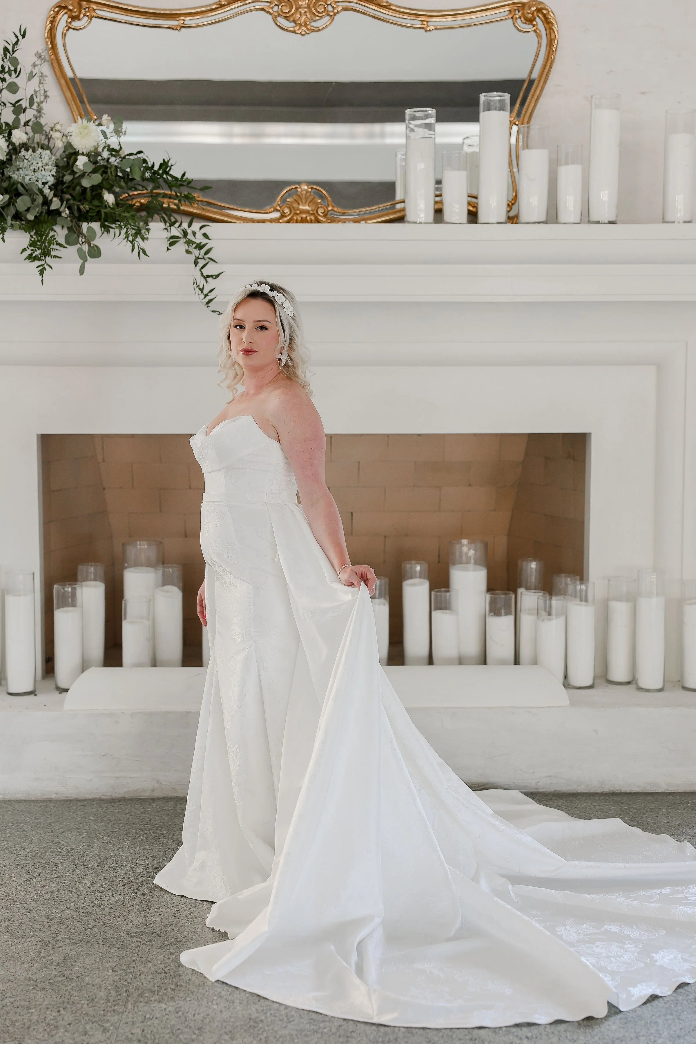 A bride wearing a strapless white wedding dress with a long train, standing in front of a white fireplace adorned with multiple white candles in glass holders. The fireplace has a decorative gold-framed mirror and a floral arrangement with greenery a