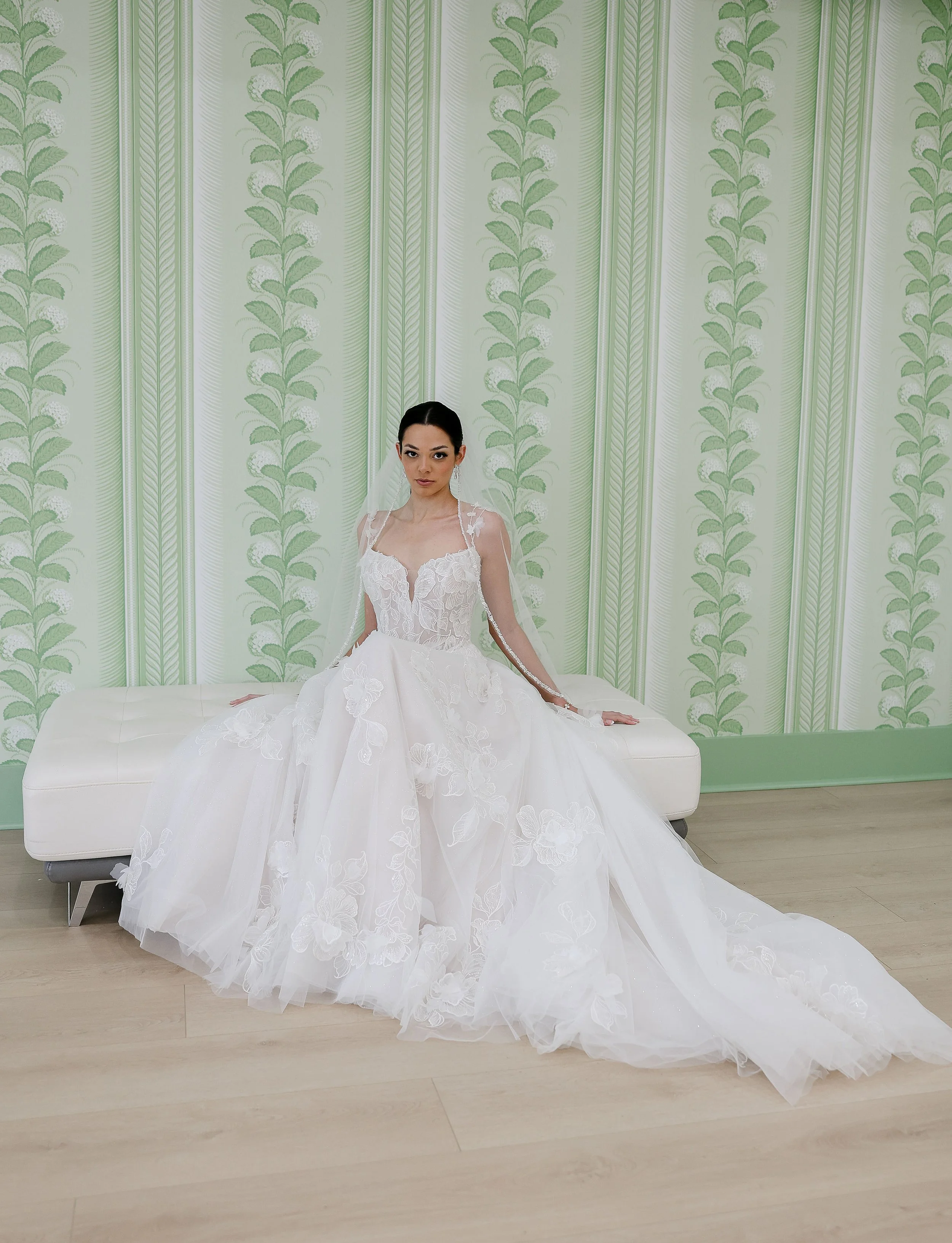 A woman in a white wedding dress sitting on a white bench in front of a green patterned wallpaper.