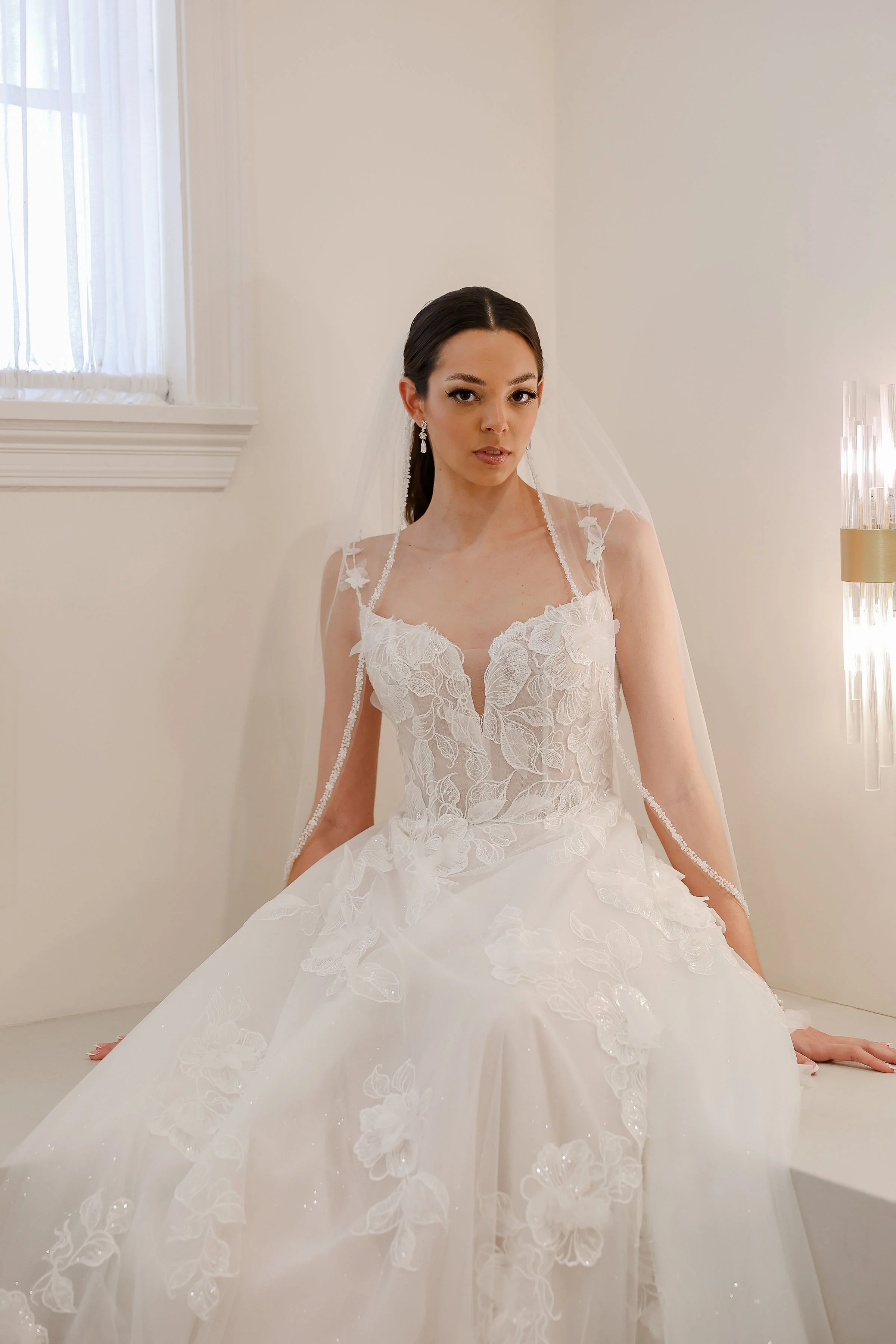 A woman in a white wedding dress sits on the floor in a room with white walls and a window with sheer curtains. She has dark hair, earrings, and a veil over her shoulders. The dress has floral lace details and a full skirt.