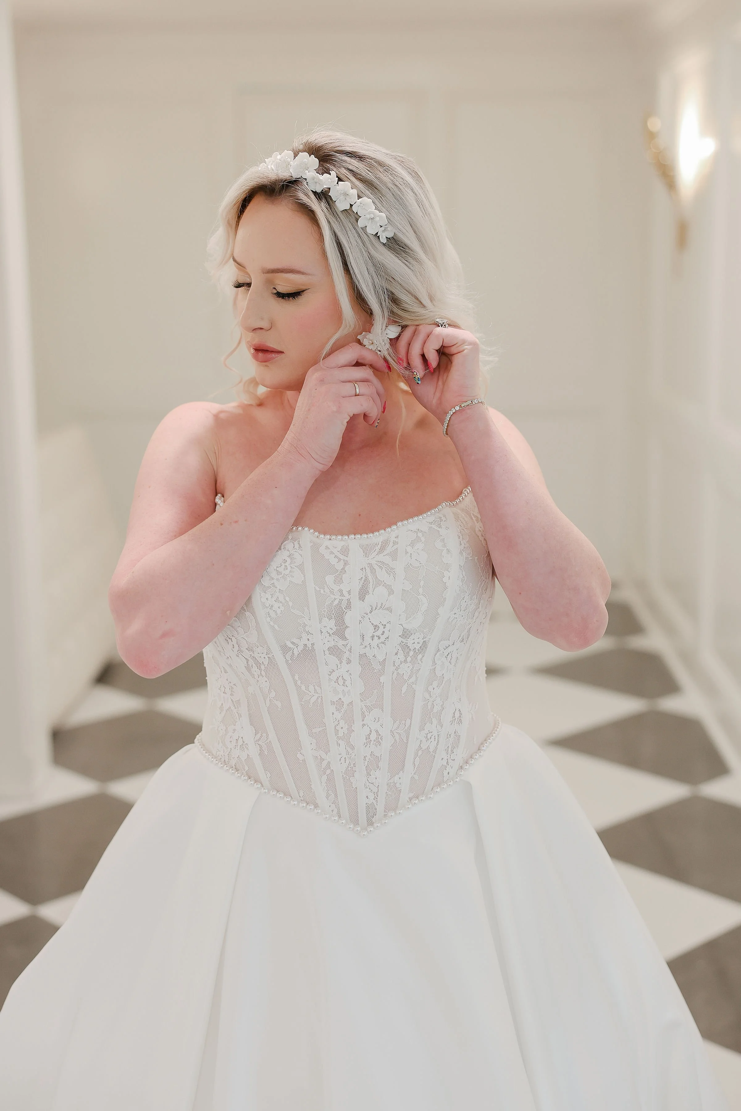 A bride with blonde hair wearing a lace wedding dress and floral headband, adjusting an earring in a room with a checkered floor.