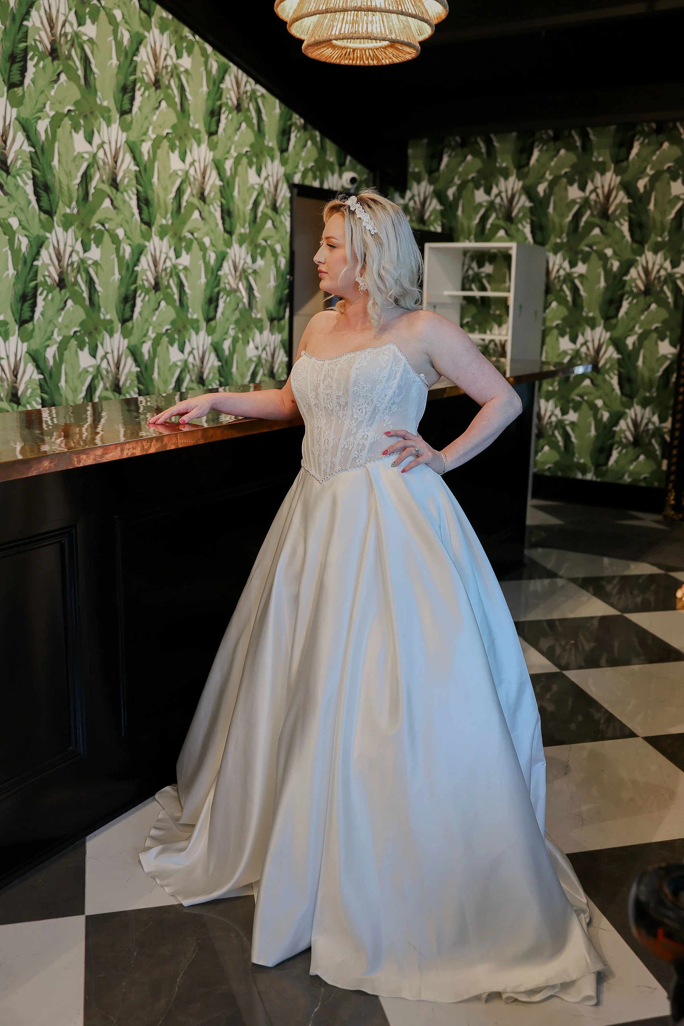 A woman in a wedding dress standing in a room with green leaf patterned wallpaper, black and white checkered marble floor, and ornate ceiling light.