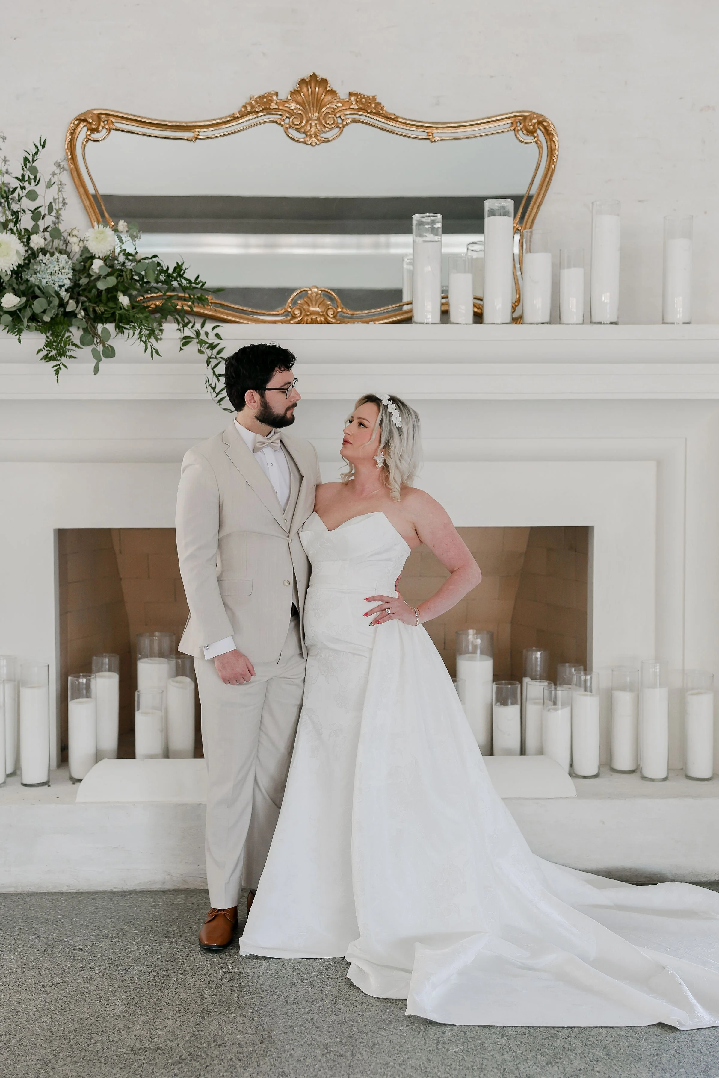 A newlywed couple standing in front of a white fireplace mantel with candles and a decorative mirror, gazing at each other. The groom is wearing a light beige suit and glasses, while the bride is in a white wedding gown with a headpiece. There is gre
