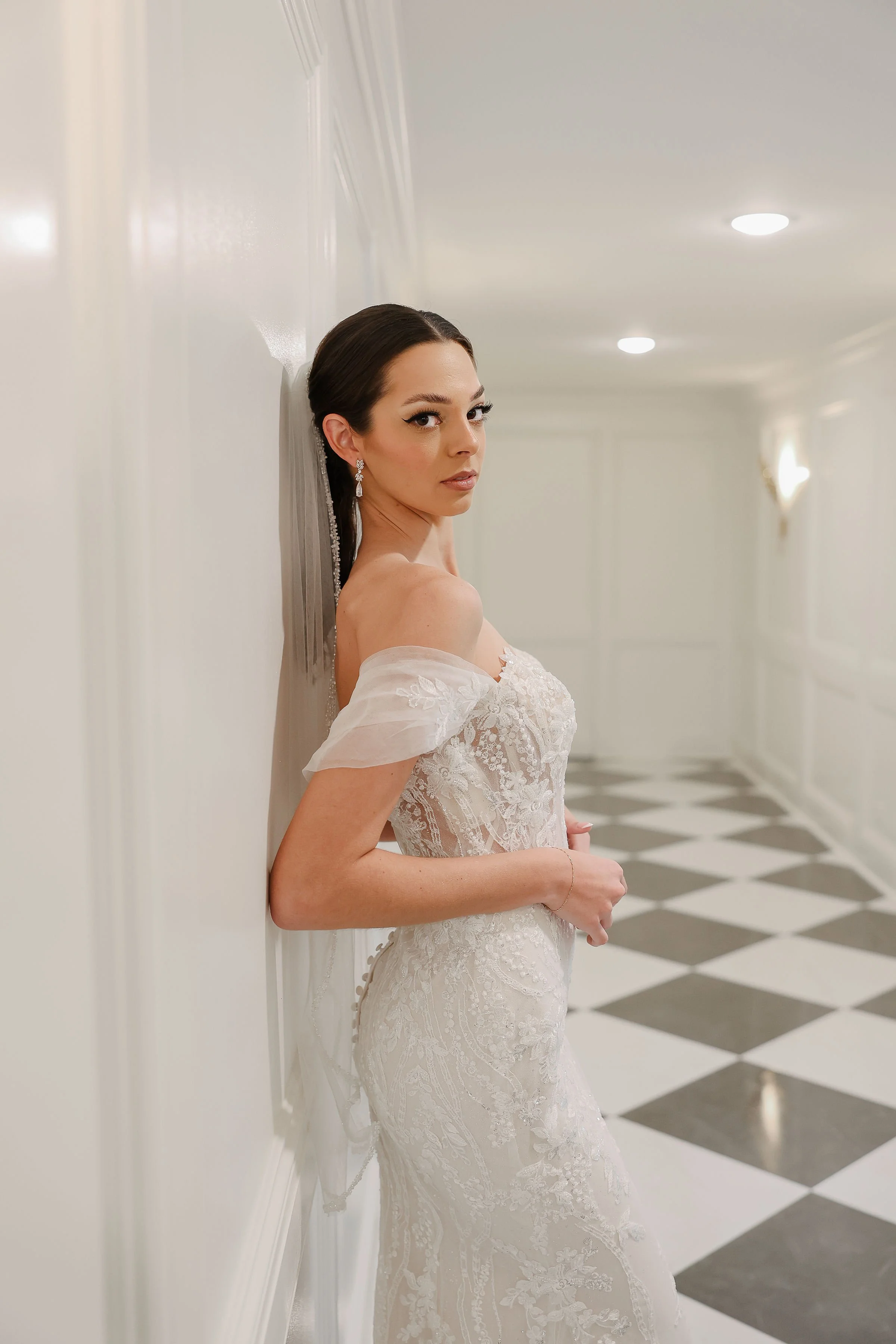 A woman in a wedding dress standing against a white wall in a hallway with black and white checkered flooring.