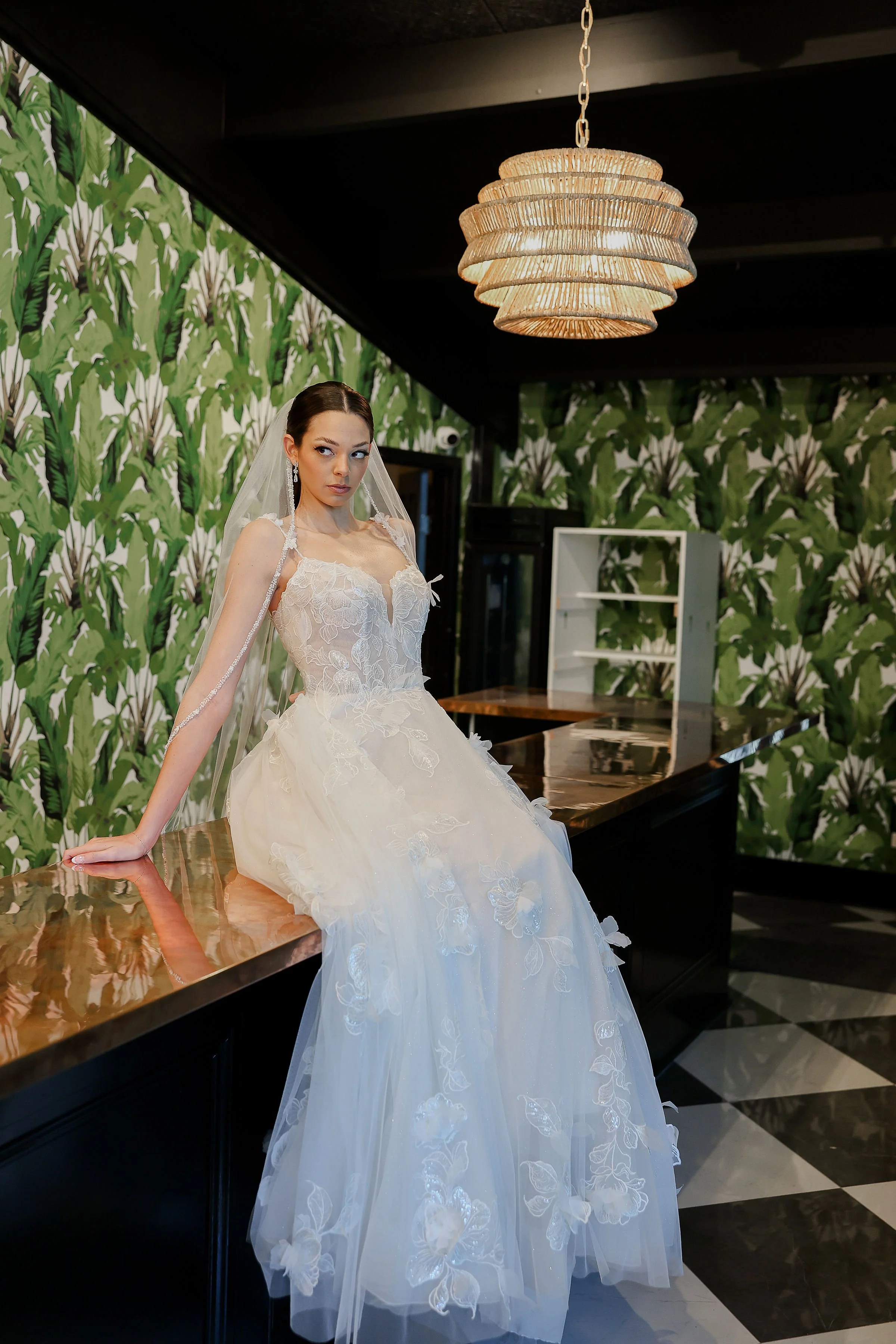 A woman in a wedding dress leaning on a table in a room with green leaf patterned wallpaper and a woven pendant light overhead.
