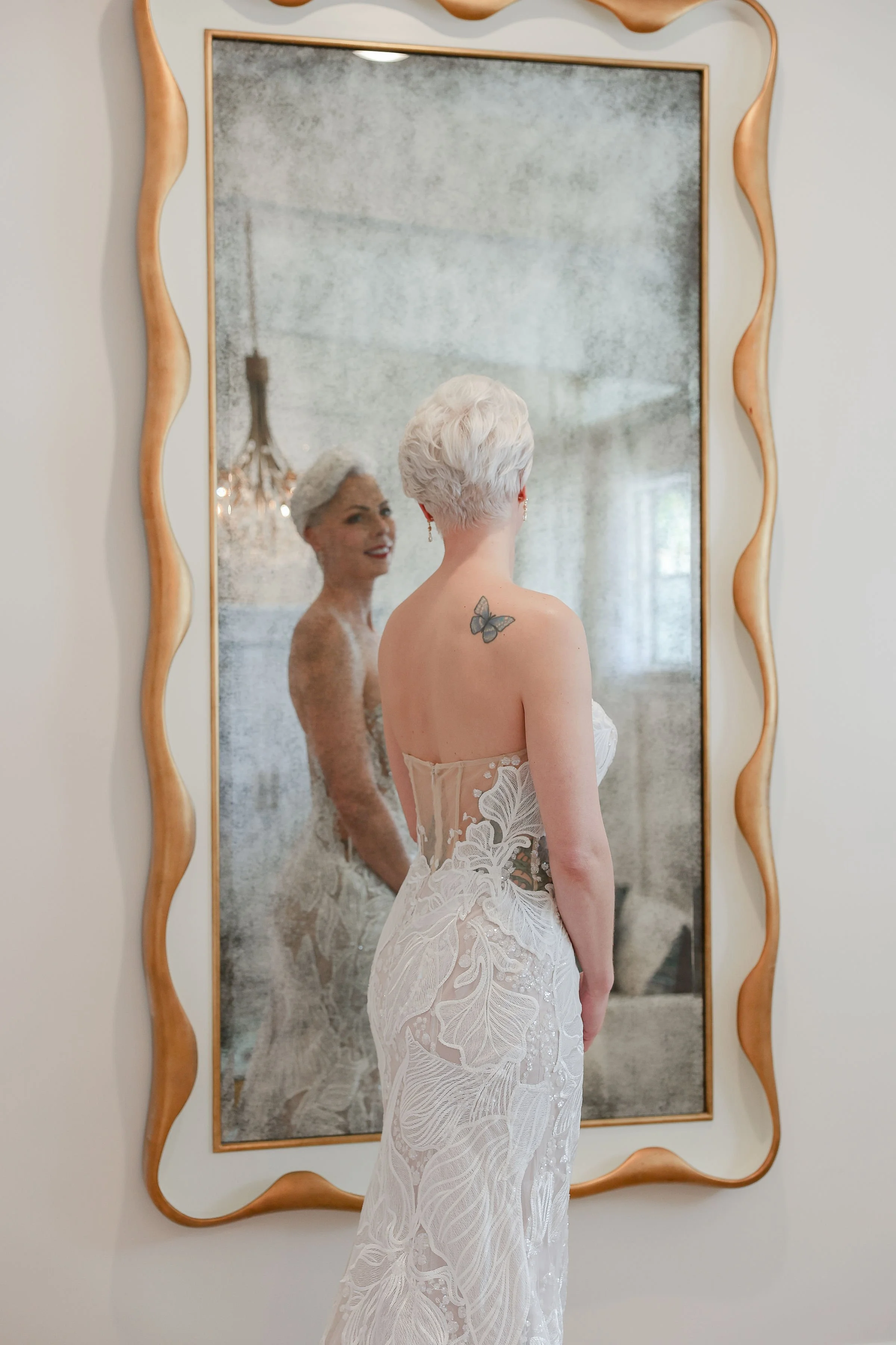 A woman with short white hair, lace wedding dress, and butterfly tattoo on her shoulder stands in front of a large ornate mirror, looking at her reflection which shows her smiling.