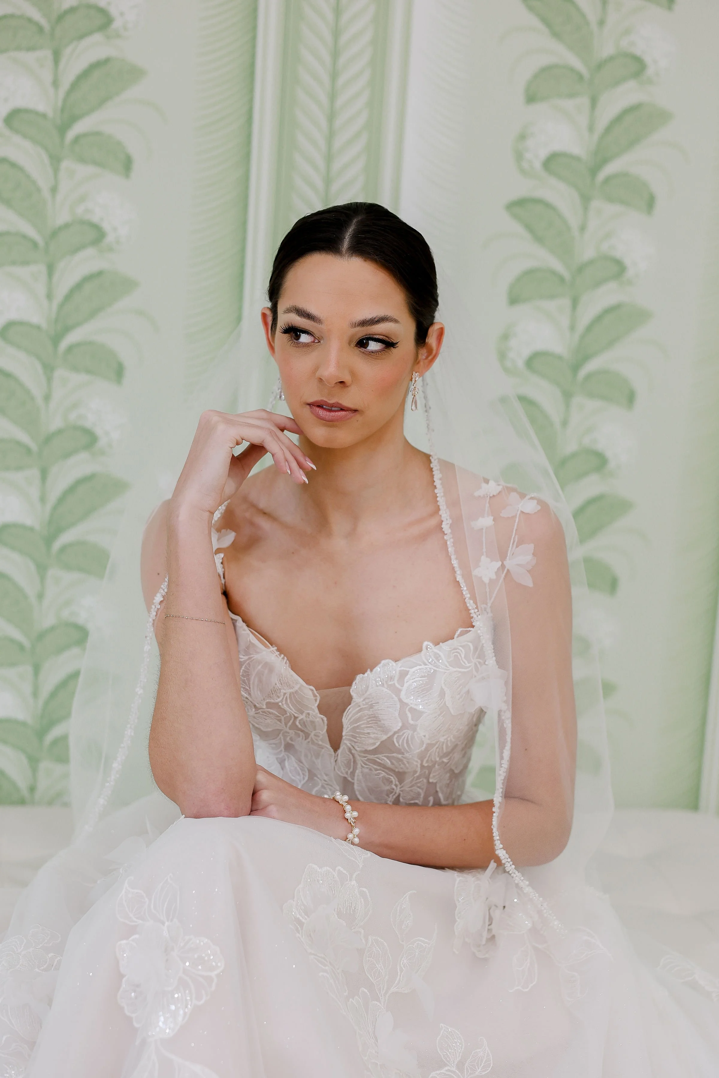 A woman with dark hair styled in an elegant updo, wearing a white wedding dress with floral embroidery, sitting in front of a green-patterned backdrop. She is touching her face gently and looking thoughtfully to the side.