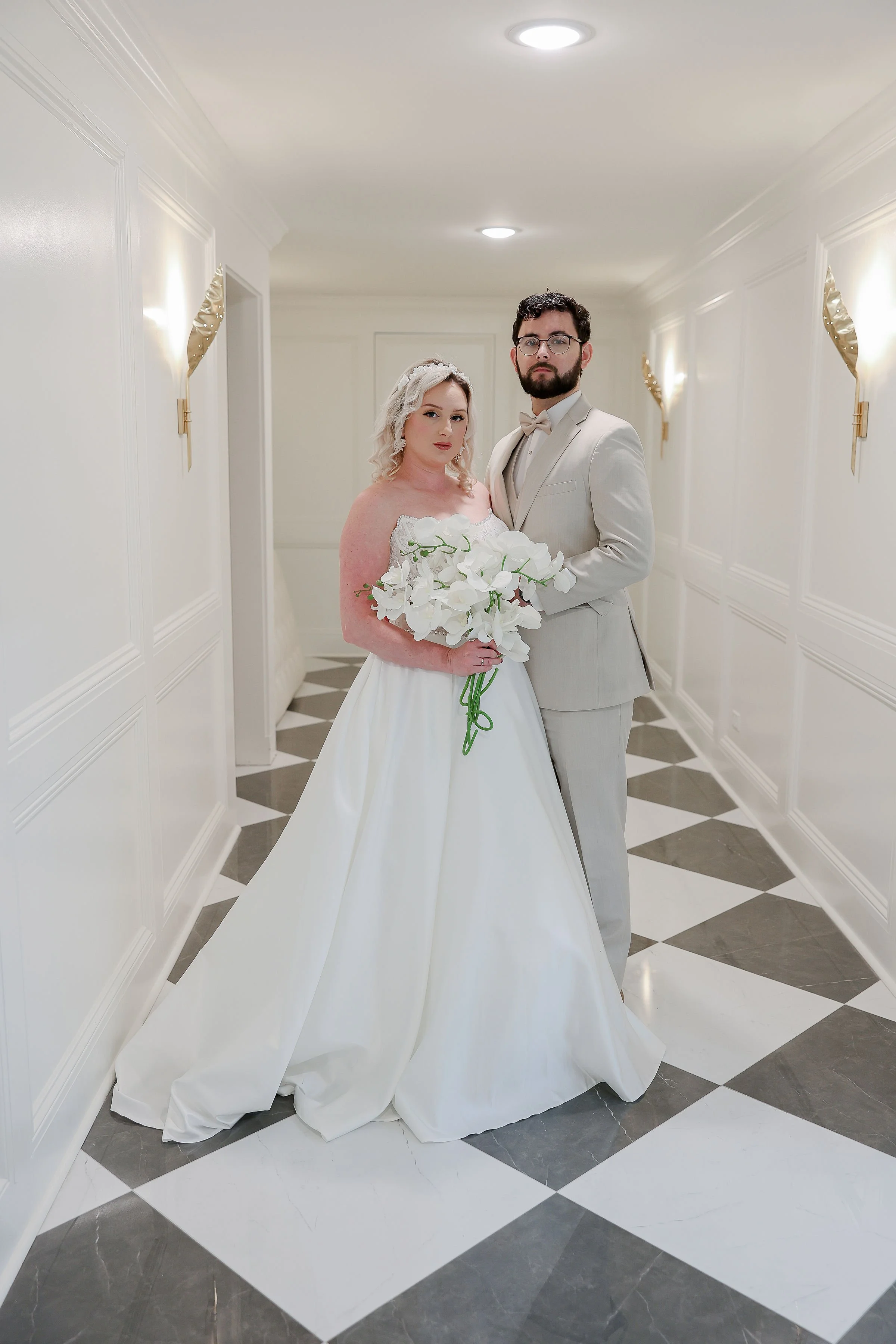 Bride and groom standing in a white, decorated hallway. The bride is holding a bouquet of white orchids, and the groom is dressed in a light suit with glasses. Elegant wall sconces are on the walls.