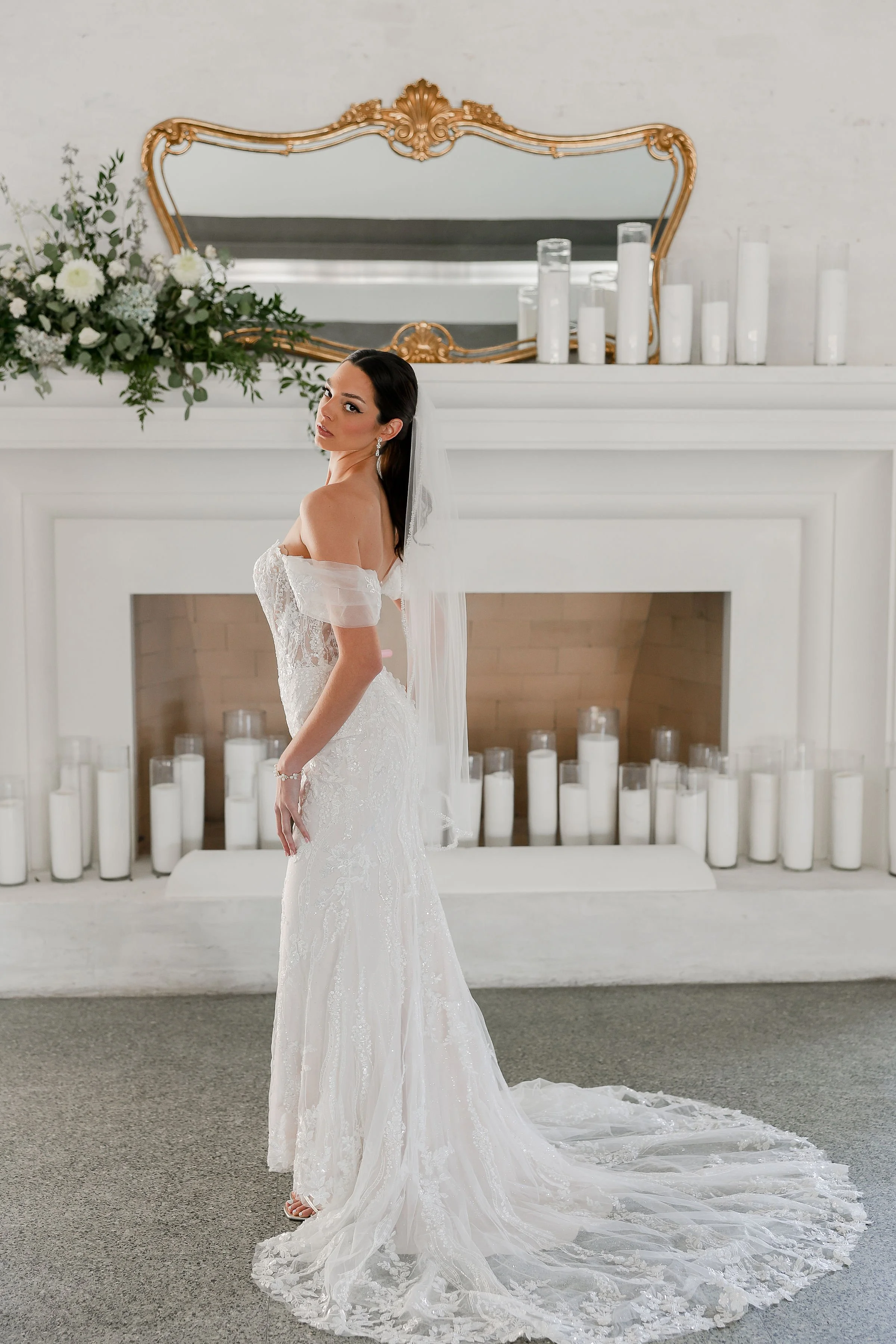 A bride stands indoors in front of a white mantelpiece decorated with white candles and an ornate gold mirror, wearing a white lace wedding gown with off-shoulder sleeves and a long train, with her hair styled back and a veil.